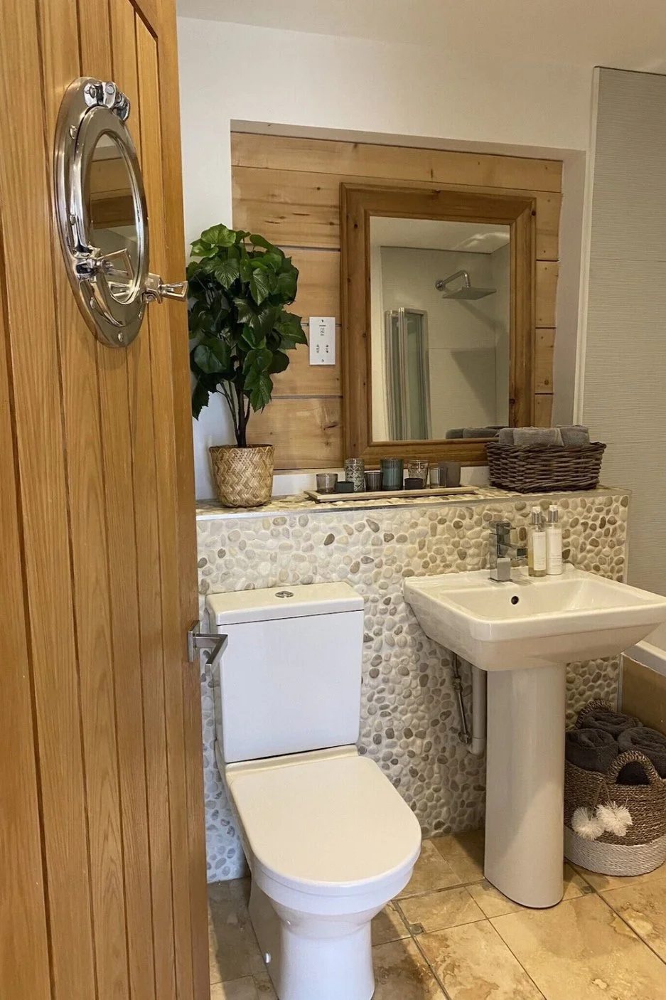 Bathroom with a wooden door, a pebble tiled wall, a small sink, a mirror, a toilet, a potted plant, candles, and baskets of towels.