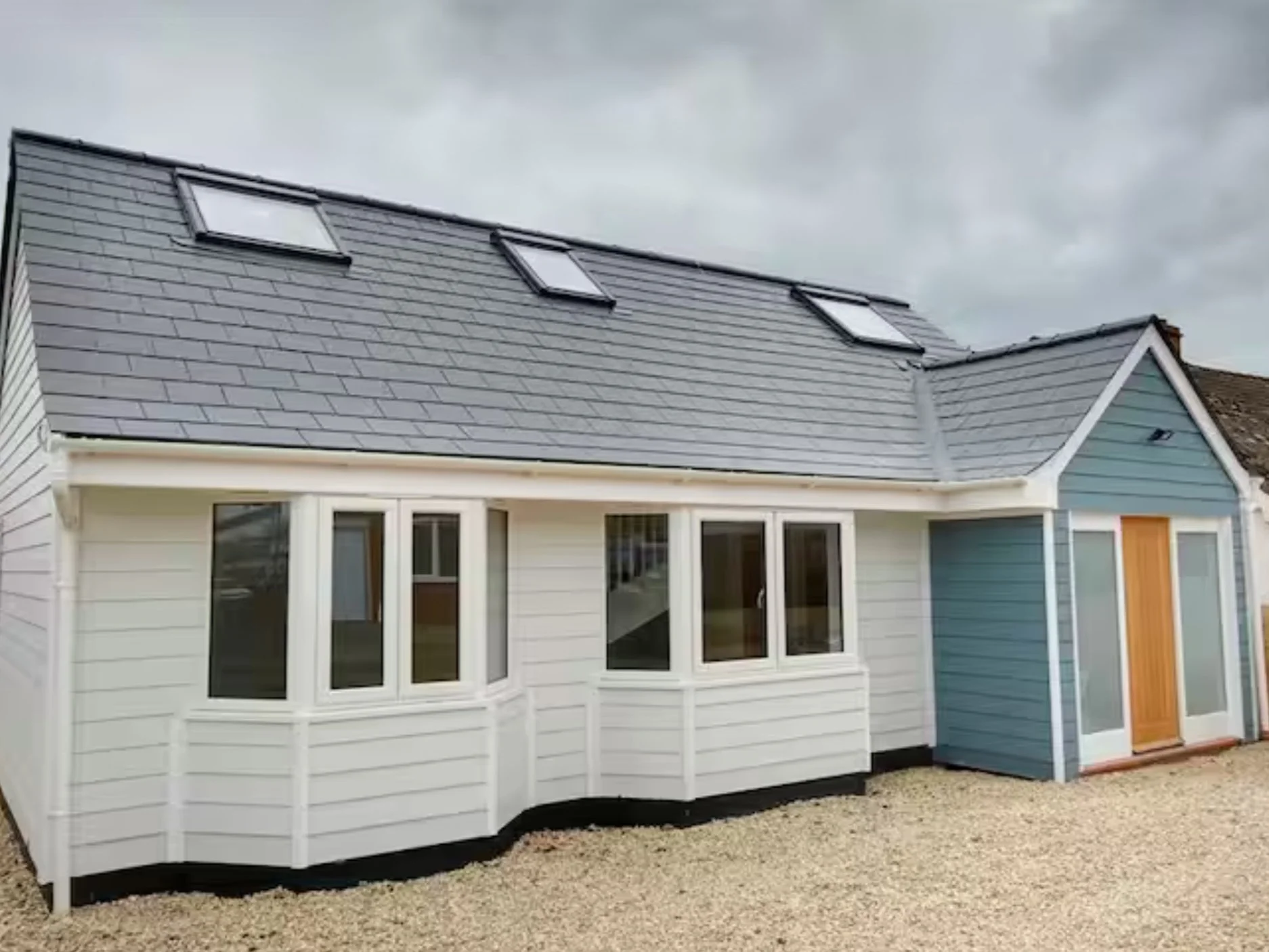 A modern cottage with a grey sloped roof featuring three skylights, white and blue exterior walls, multiple windows, and a gravel yard under a cloudy sky.
