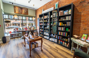 Inside the Serendipity Bookstore in Chelsea Mi, the front section of the store with bookshelves against a brick wall, wooden table in the center of the room, and the front windows looking out onto Main St, Chelsea.