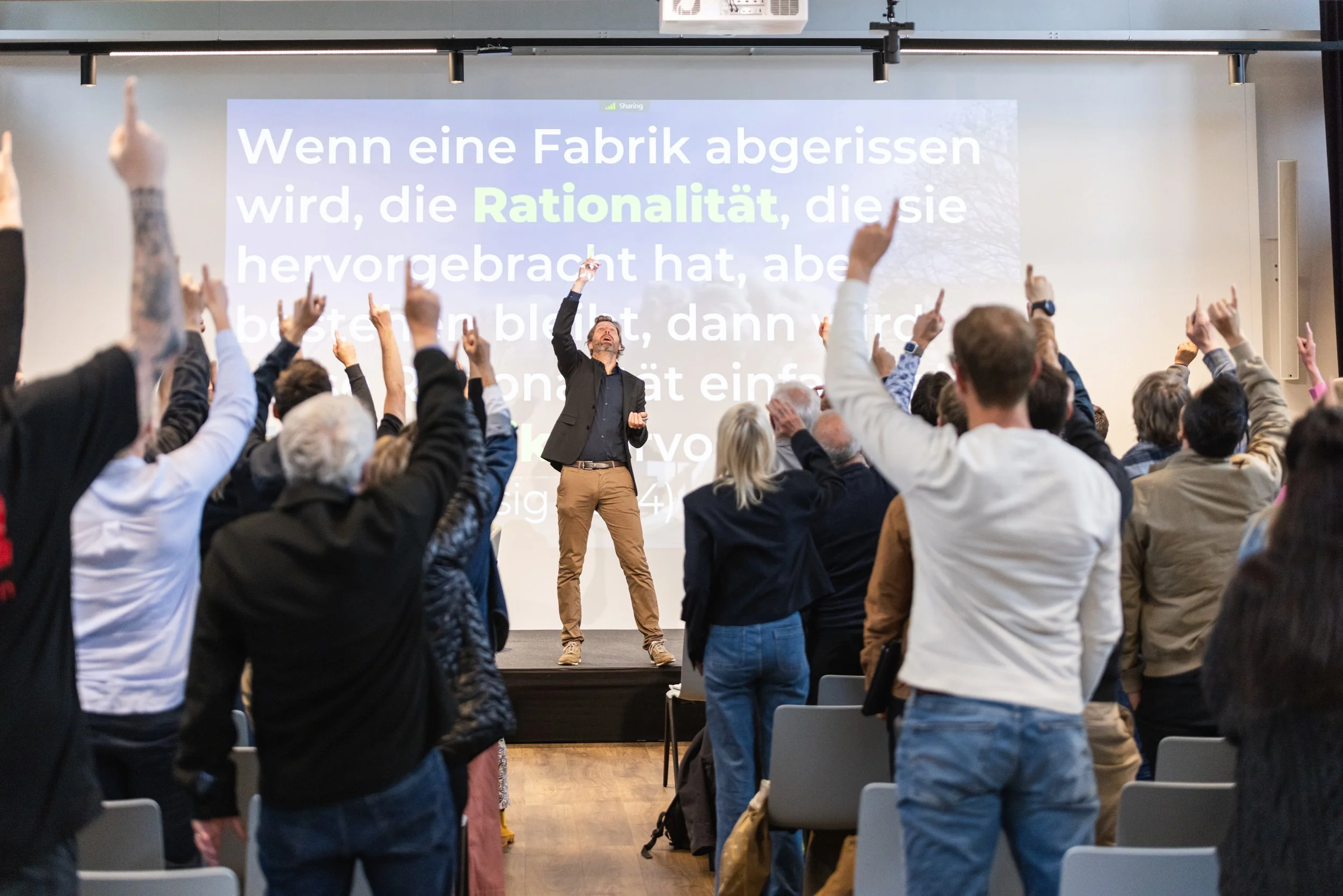 A man in a dark suit and tan pants giving a presentation in front of a large screen, with an engaged audience raising their hands in the conference room.