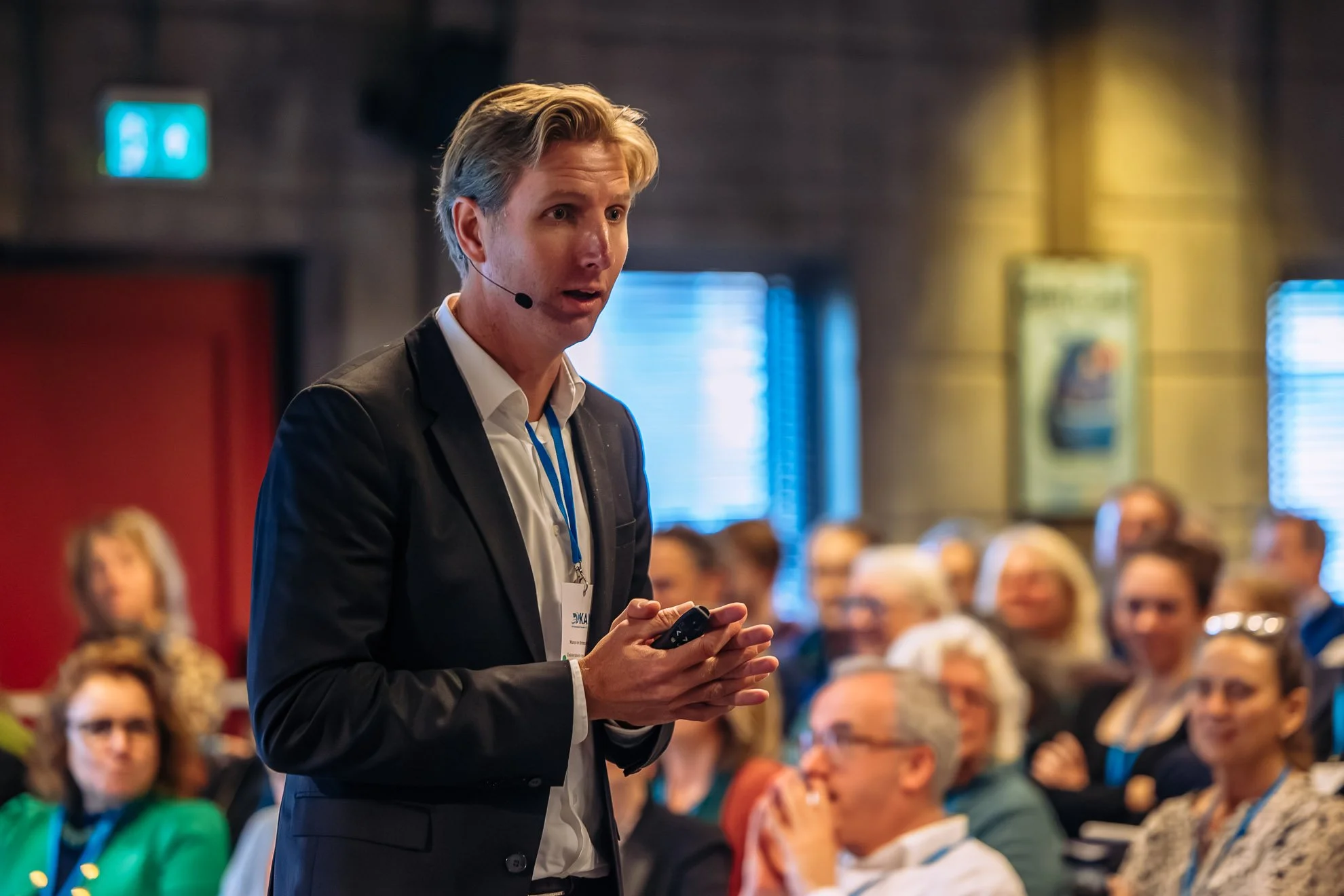 A man with blonde hair, wearing a dark suit and white shirt, is holding a microphone and a small device while speaking at a conference or seminar. He is standing in front of a seated audience in a dimly lit room with a blurred background of people and screens.