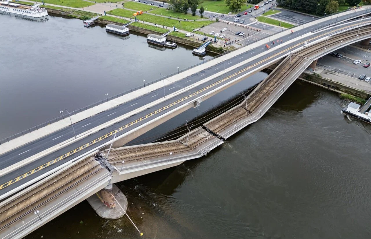 Aerial view of a bridge over a river with rail tracks underneath, cars on the bridge, and a park with boats docked along the water nearby.