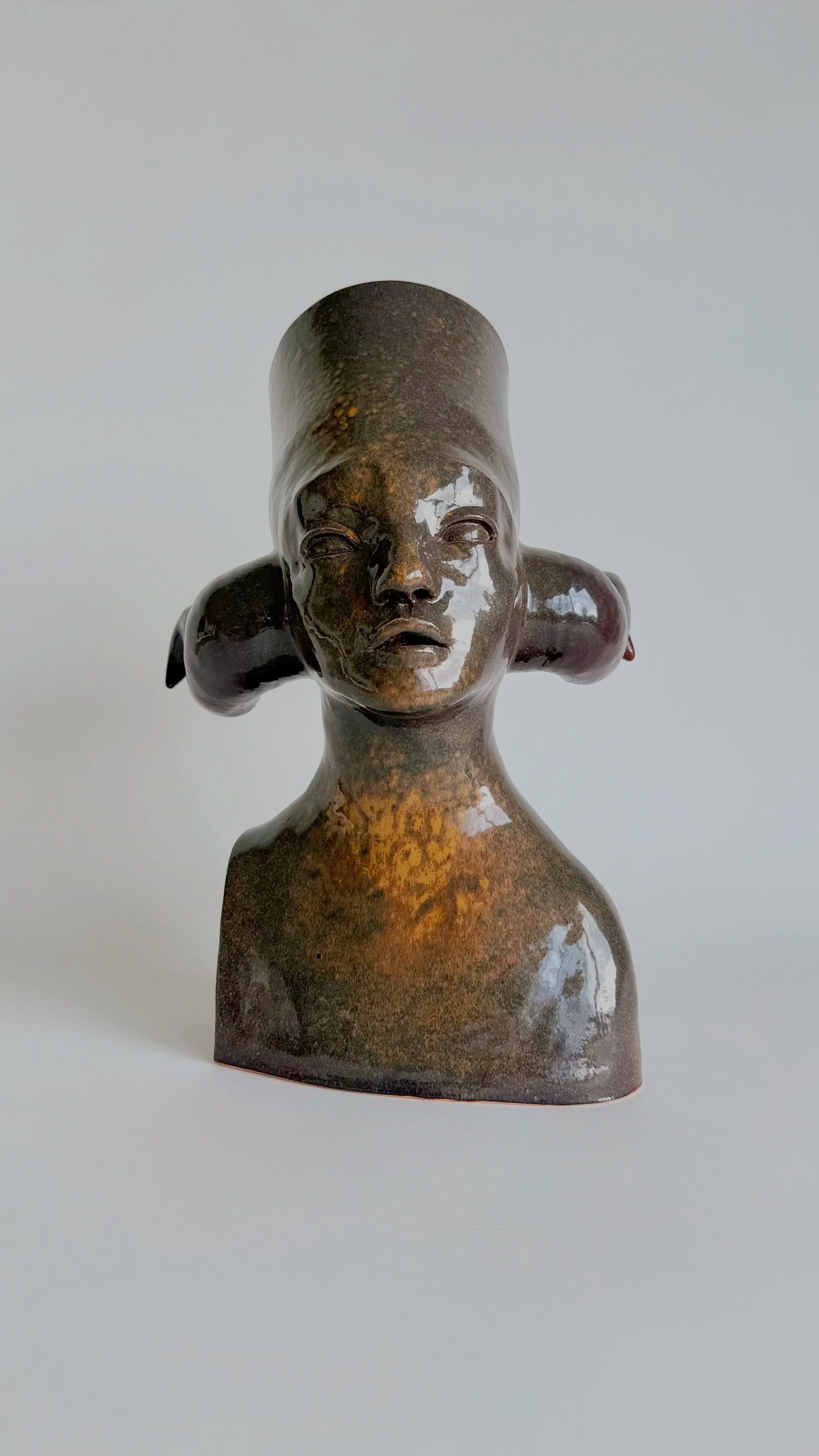 A ceramic sculpture by Mateo Aranda. Bust of a human-like creature with horns and head-piece. Stoneware, brown-golden glaze.