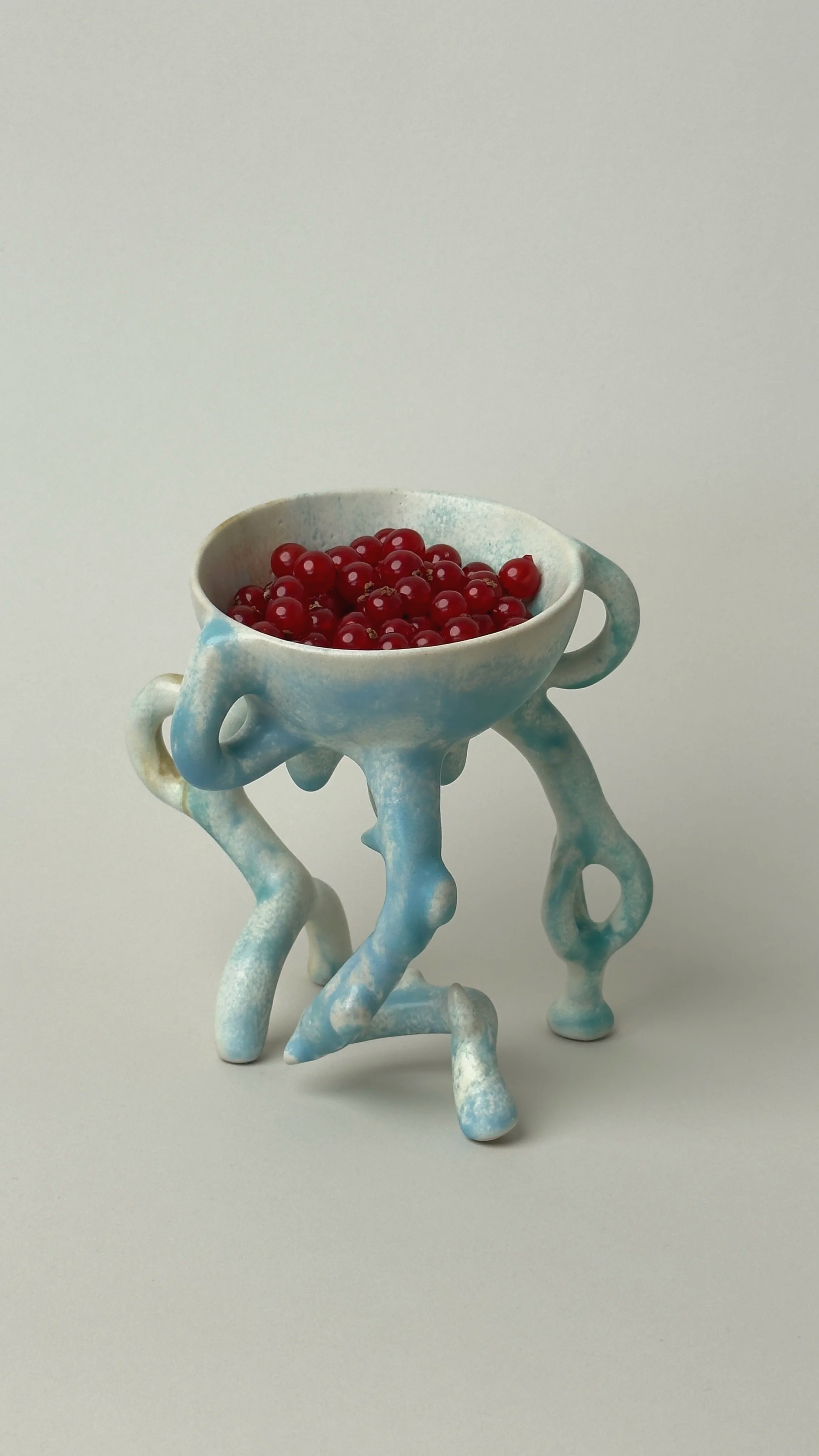 A ceramic bowl filled with red currants placed on an artistic, abstract ceramic stand with multiple looping and twisting limbs, set against a plain white background.