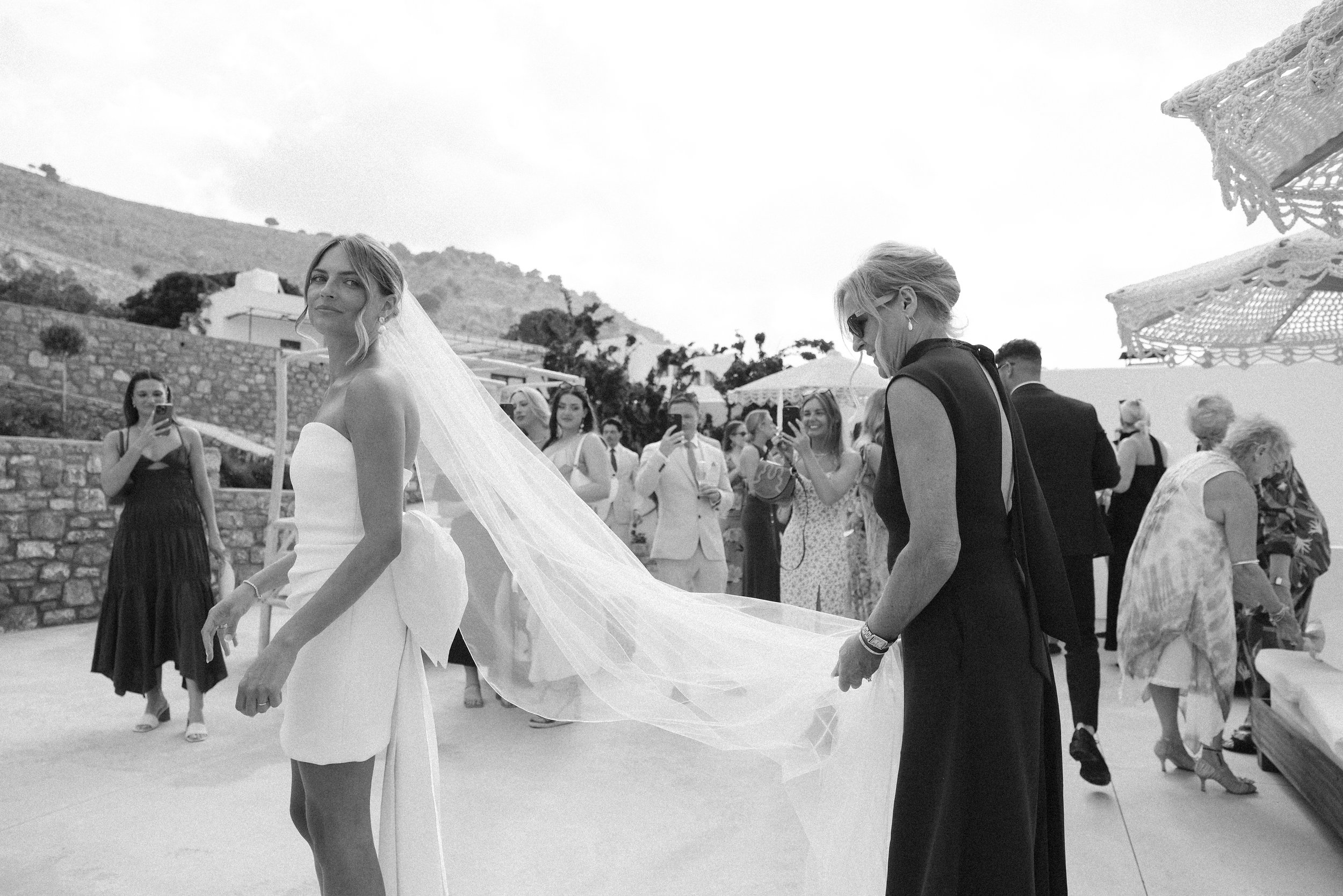 A bride in a wedding dress with a long veil, standing outdoors at a wedding reception, surrounded by guests taking photos. Hills and stone walls are in the background.
