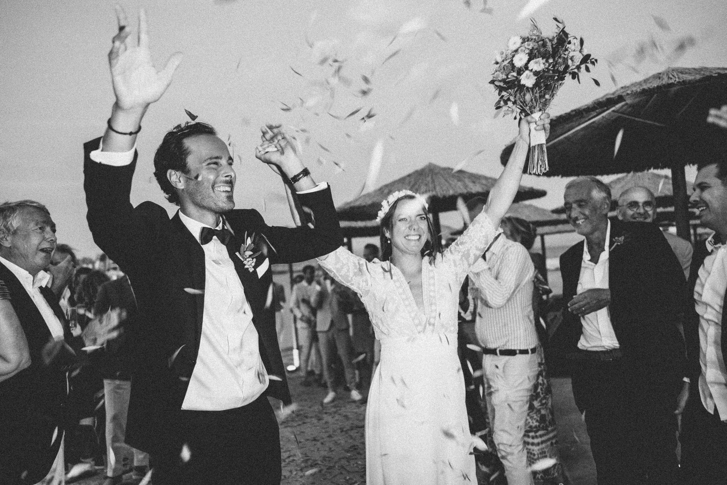 A black-and-white photo of a joyful wedding celebration on a beach. The bride and groom are smiling, raising their hands, and surrounded by guests. The bride is holding a bouquet of flowers.