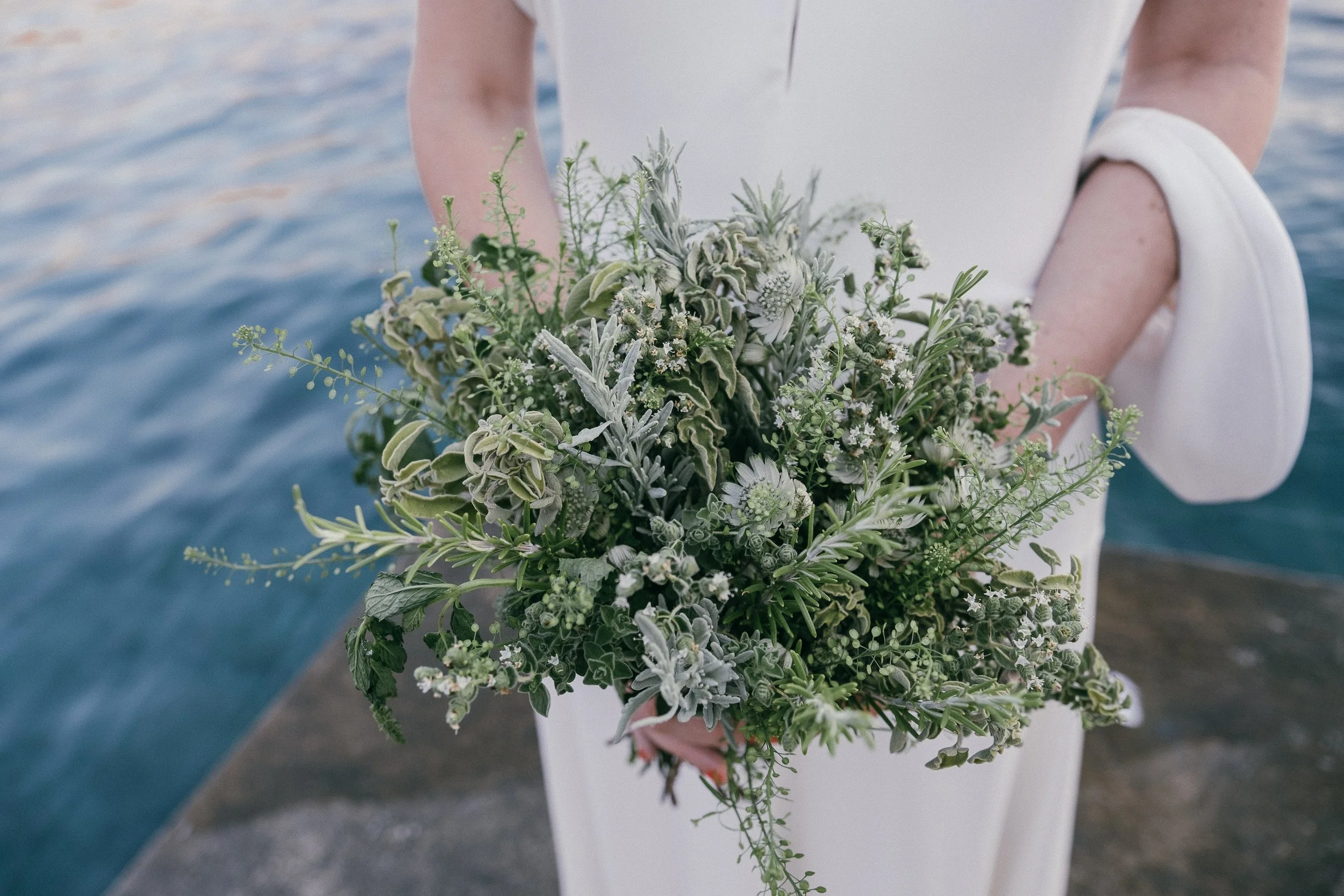 A woman in a white dress holds a bouquet of green and white flowers, with a body of water in the background.