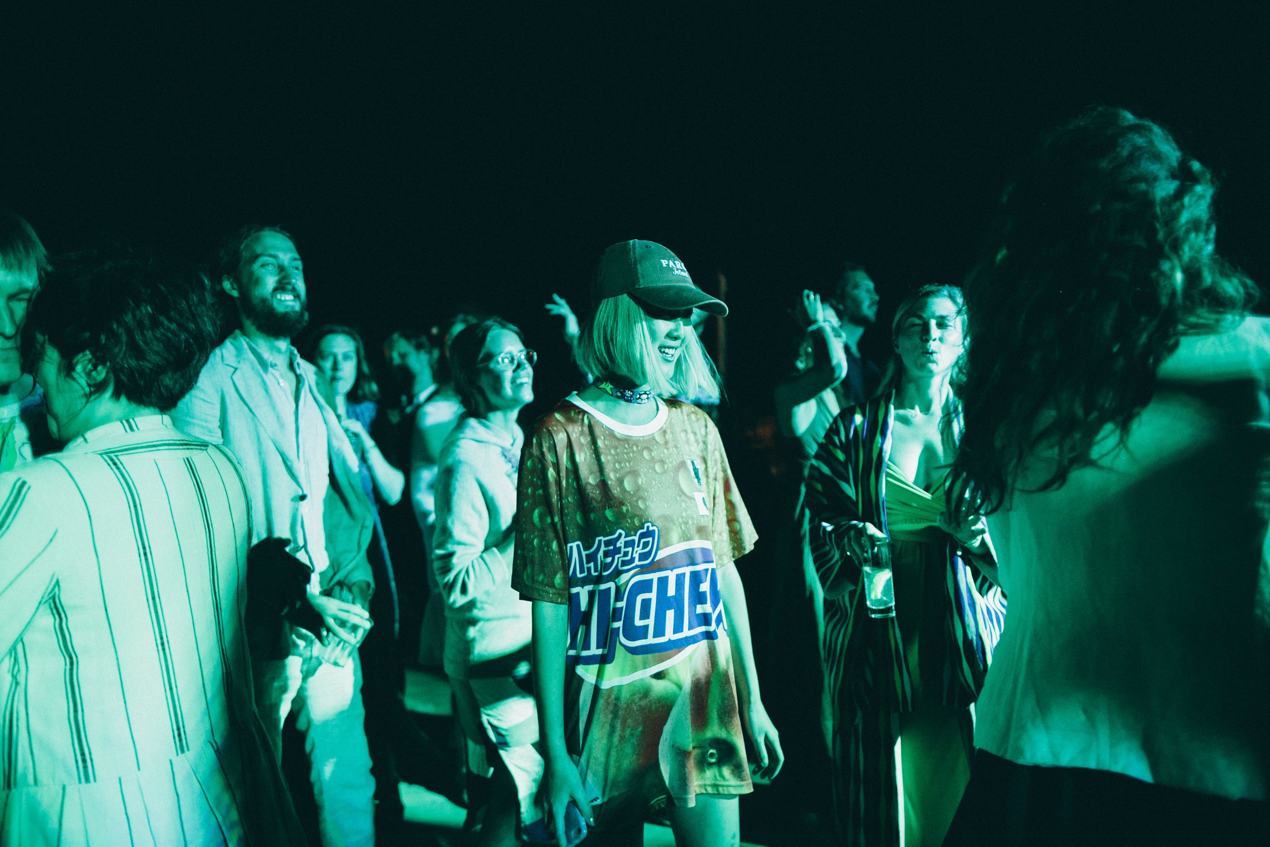 Group of people enjoying a concert or event, illuminated by green stage lighting, with some dancing and others talking, in a dark environment.