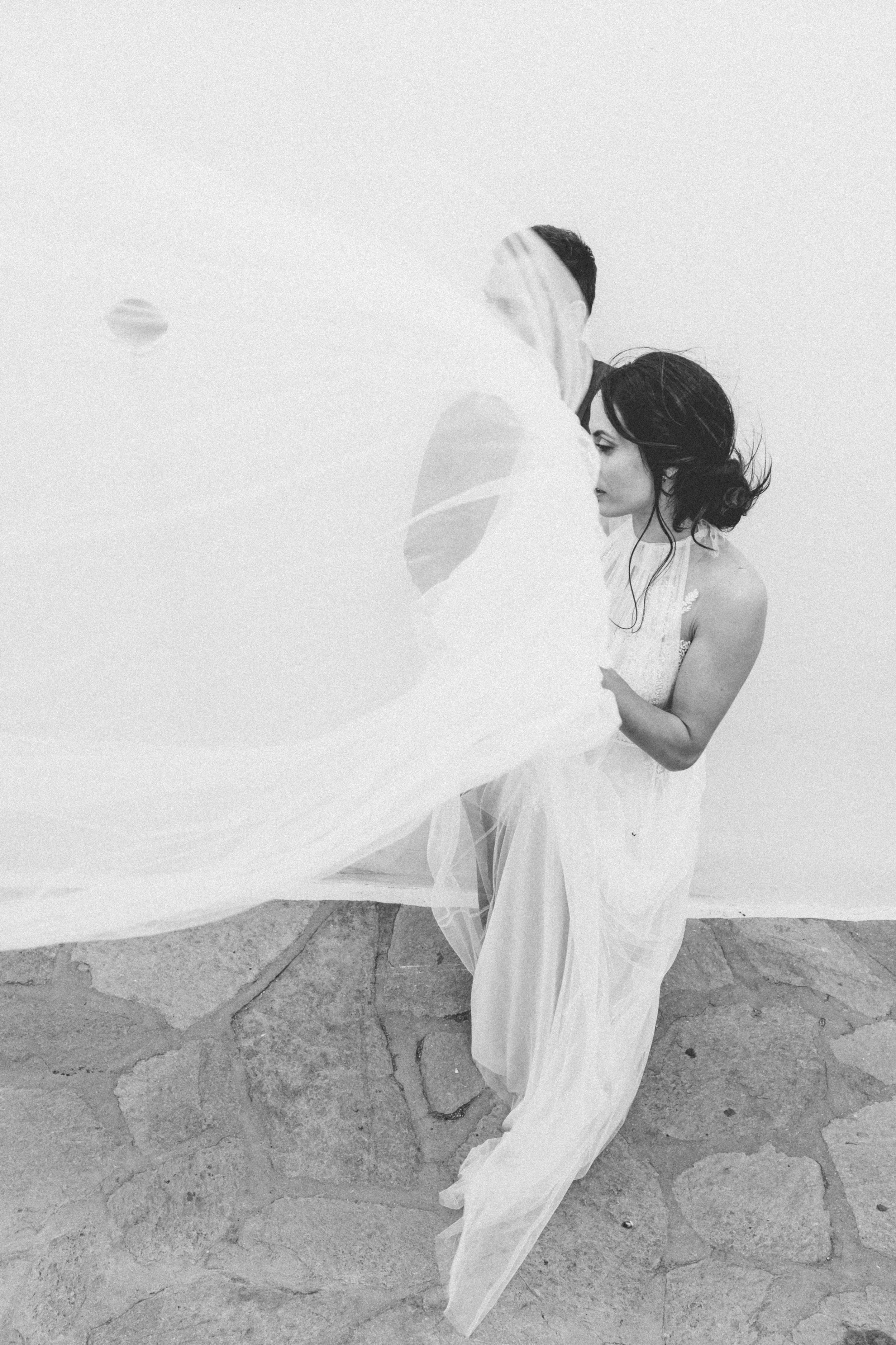 Black and white photo of a man and woman embracing, with the woman wearing a flowing dress and the man partially obscured by fabric in the foreground, standing on a stone surface against a plain white background.