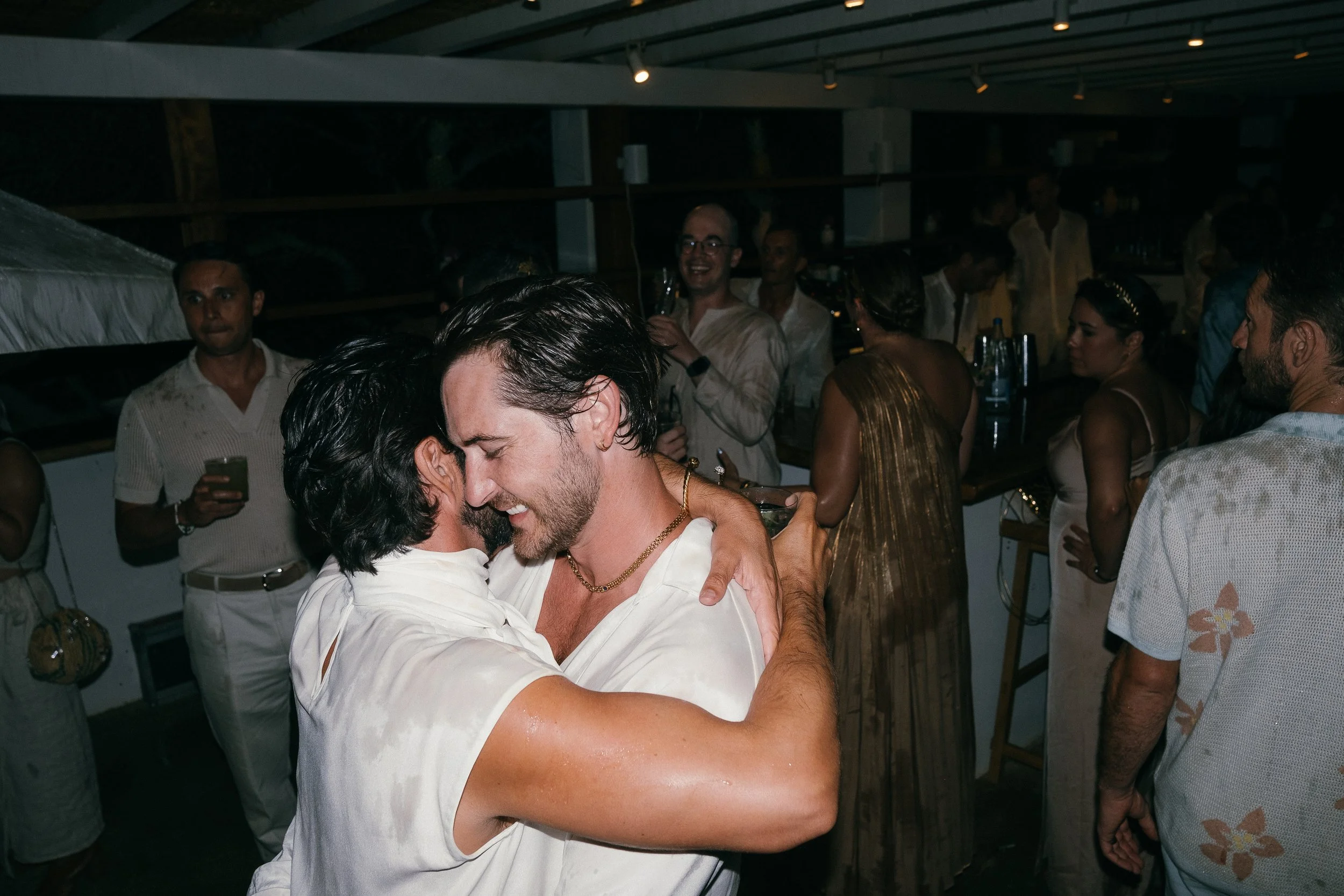 Two men embracing and dancing at a party with other guests in the background.