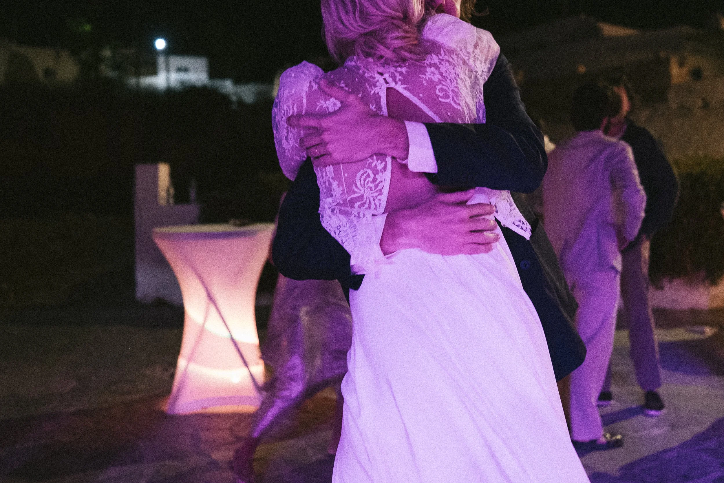 A person in a white lace dress hugging another person at a nighttime outdoor event, with other people and a lit high table in the background.