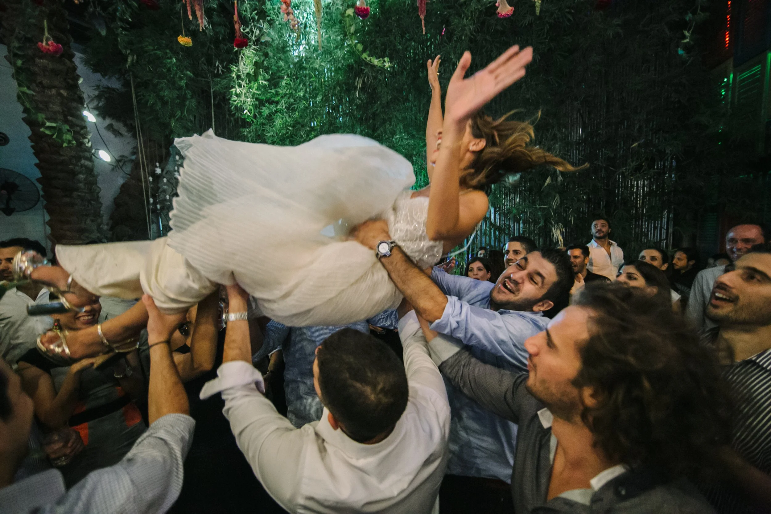 Group of people celebrating, lifting a woman in a white dress during a lively event at night.