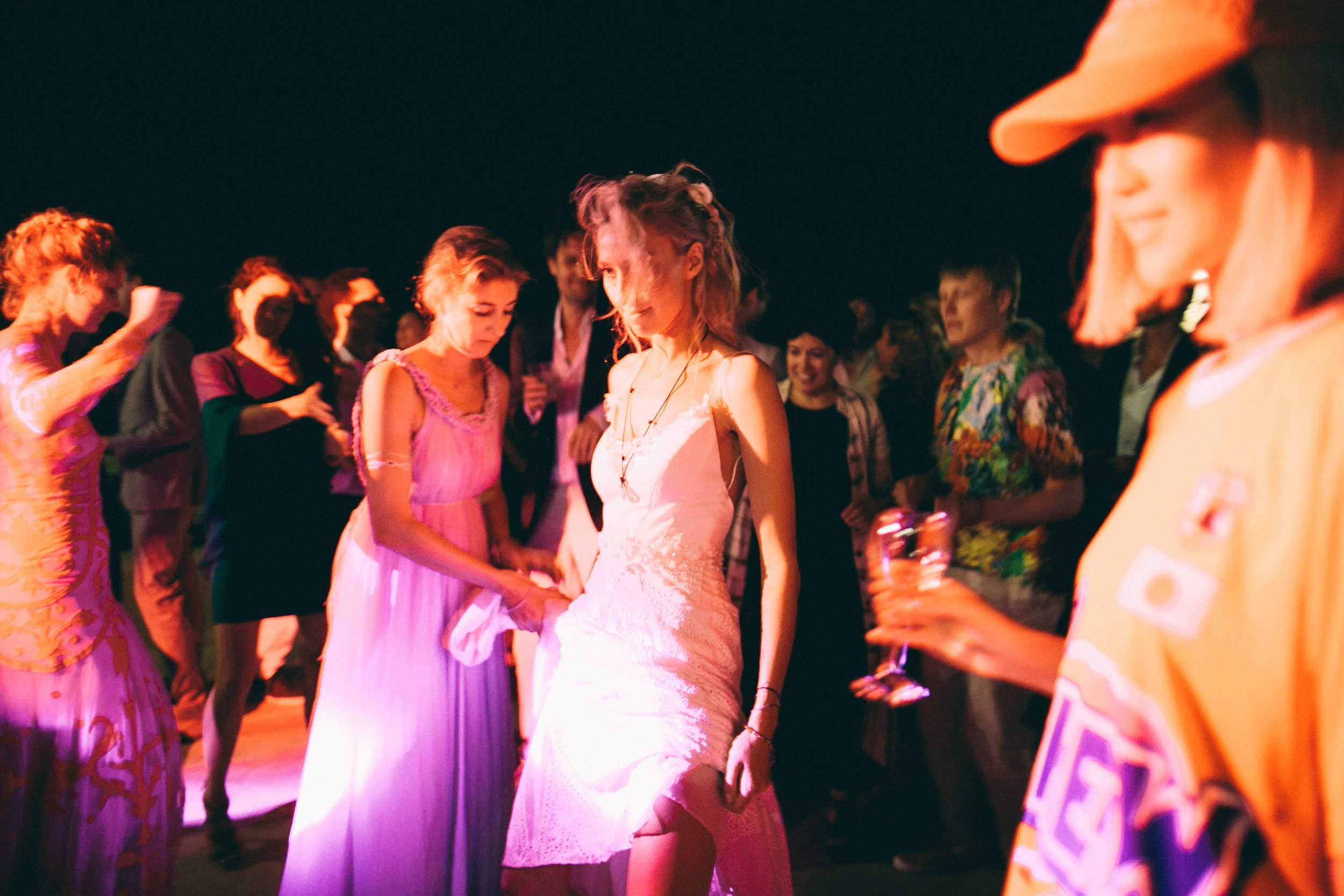 People dancing at a party or celebration, with a woman in a white dress in the center, surrounded by others in colorful outfits, under warm lighting.