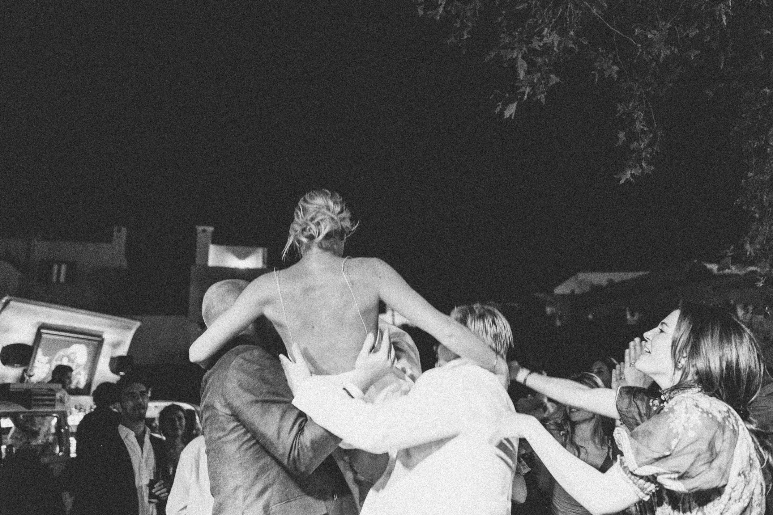 People celebrating at a nighttime outdoor event, lifting a woman on a person's shoulders, as others reach out to her, with a large screen and trees in the background.