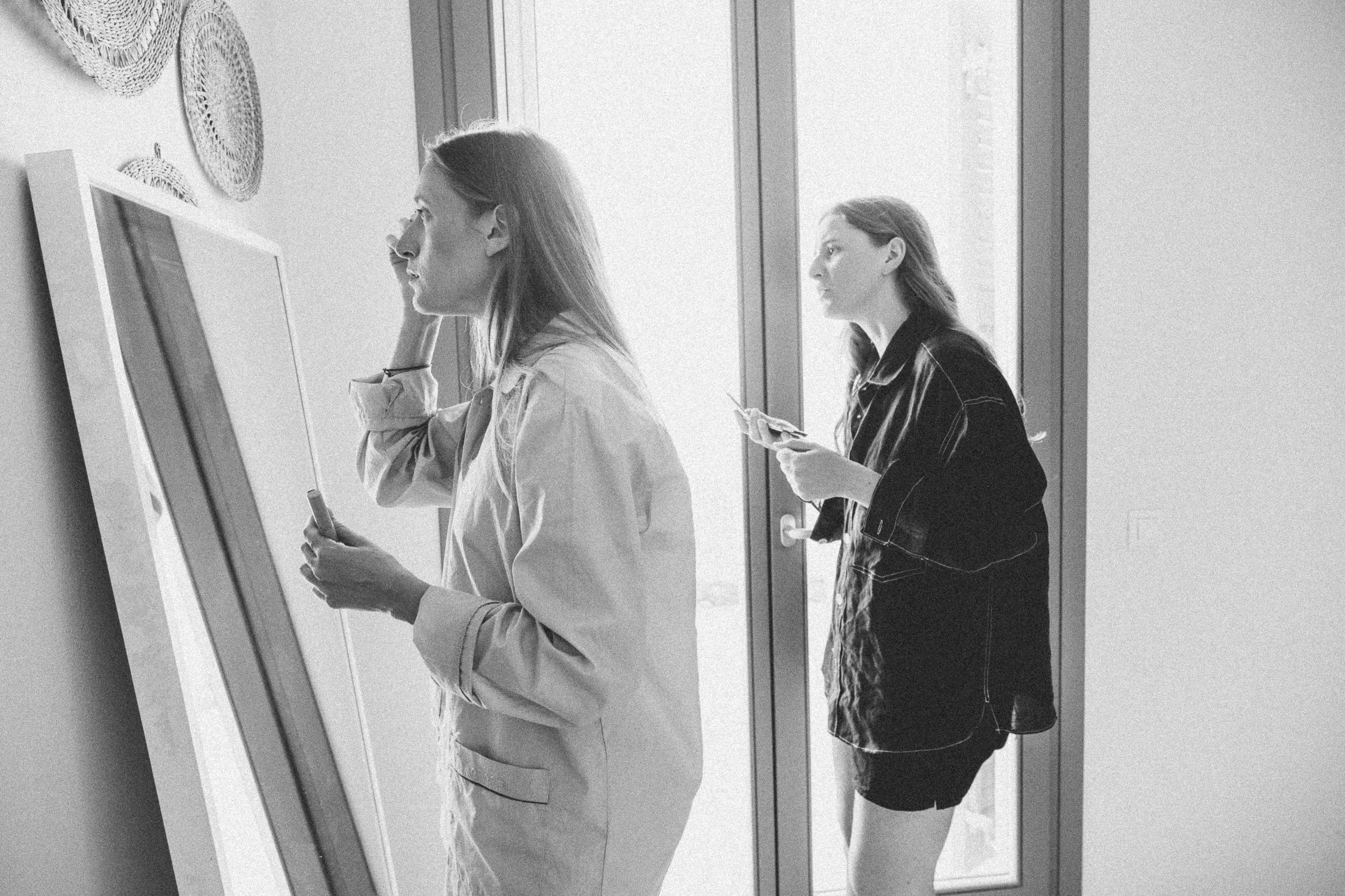 Two young women standing near a large mirror in a well-lit room. One woman is looking at her reflection with a serious expression, while the other woman is standing behind her, looking at her phone. The room has a sliding glass door and decorative wa