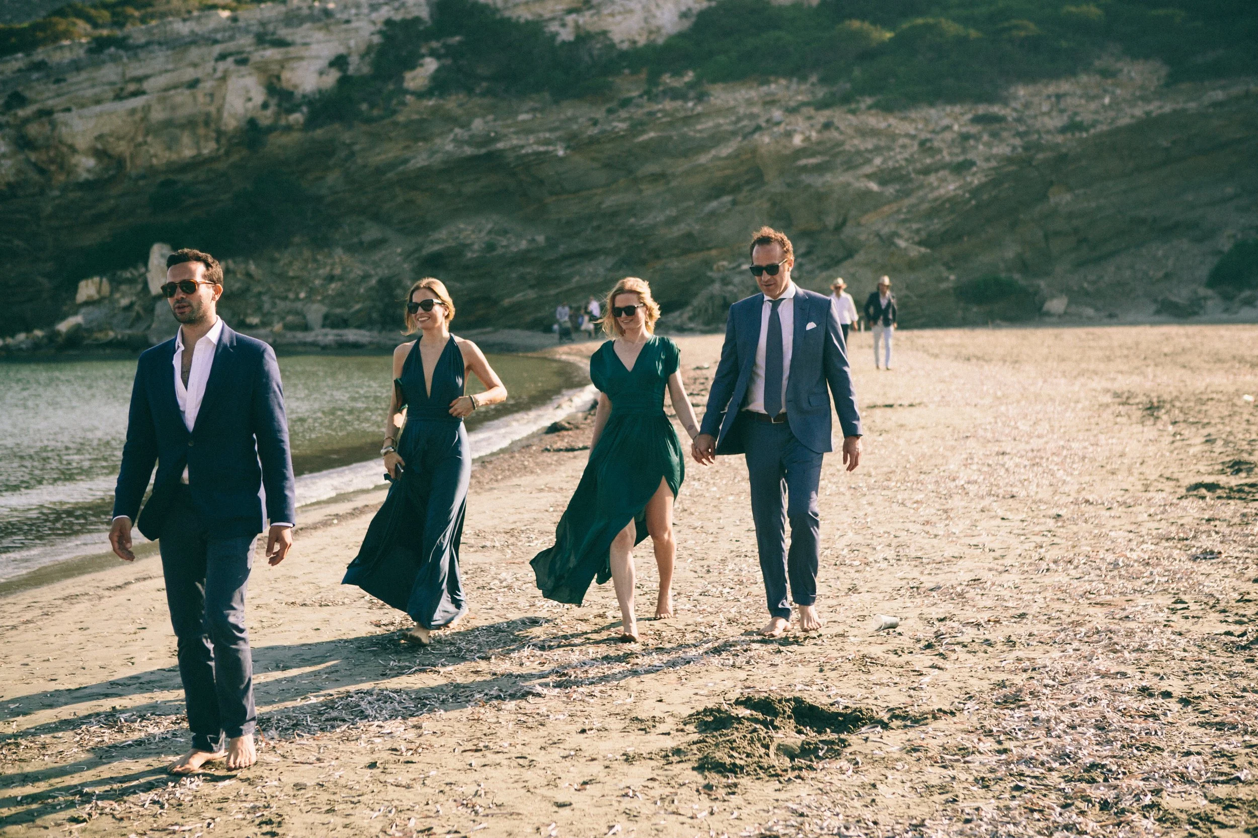 Group of adults in formal attire walking barefoot along a sandy beach, with a rocky hillslope in the background.