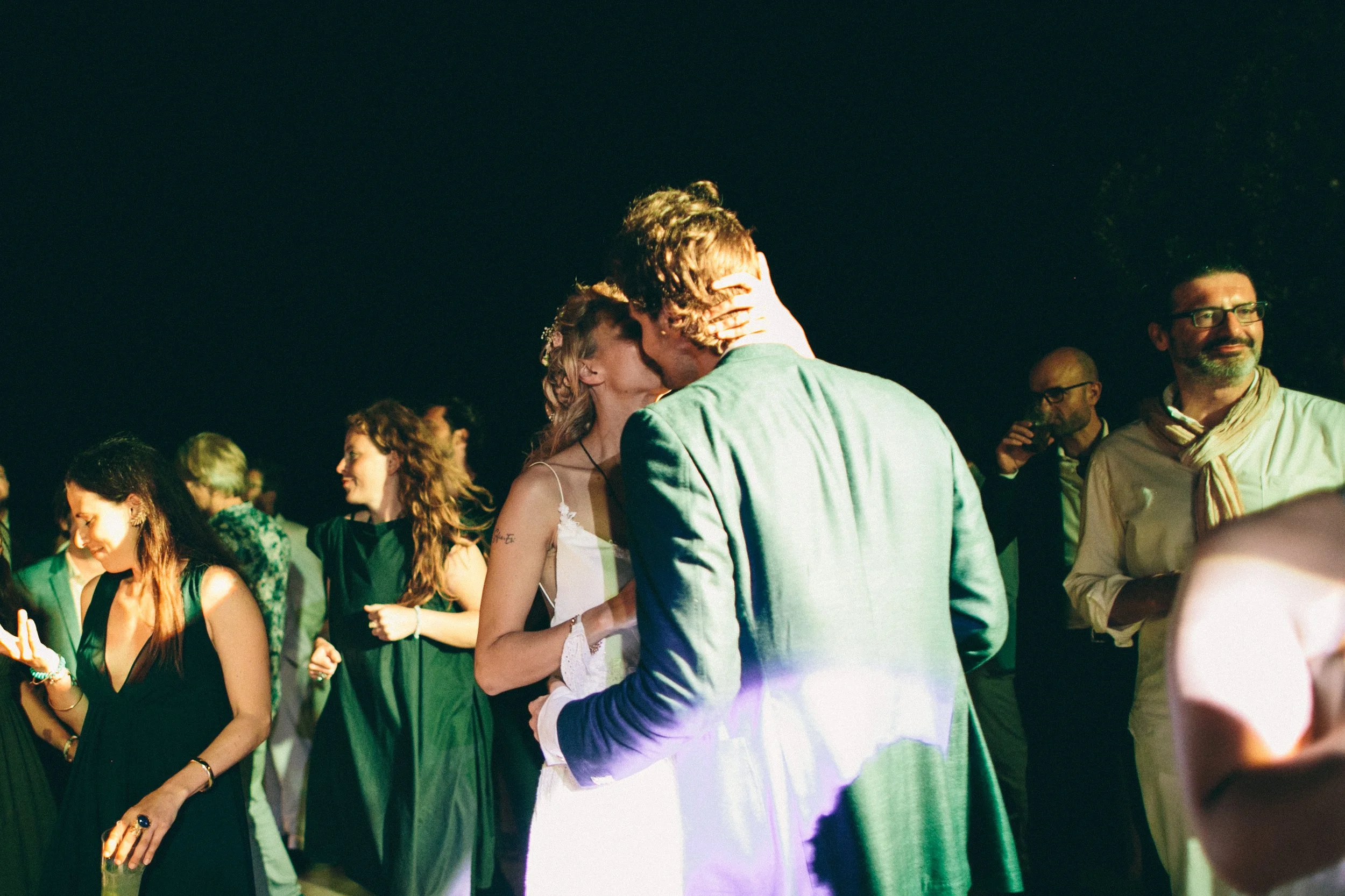 A couple sharing a kiss at night during a social gathering, surrounded by other people socializing.