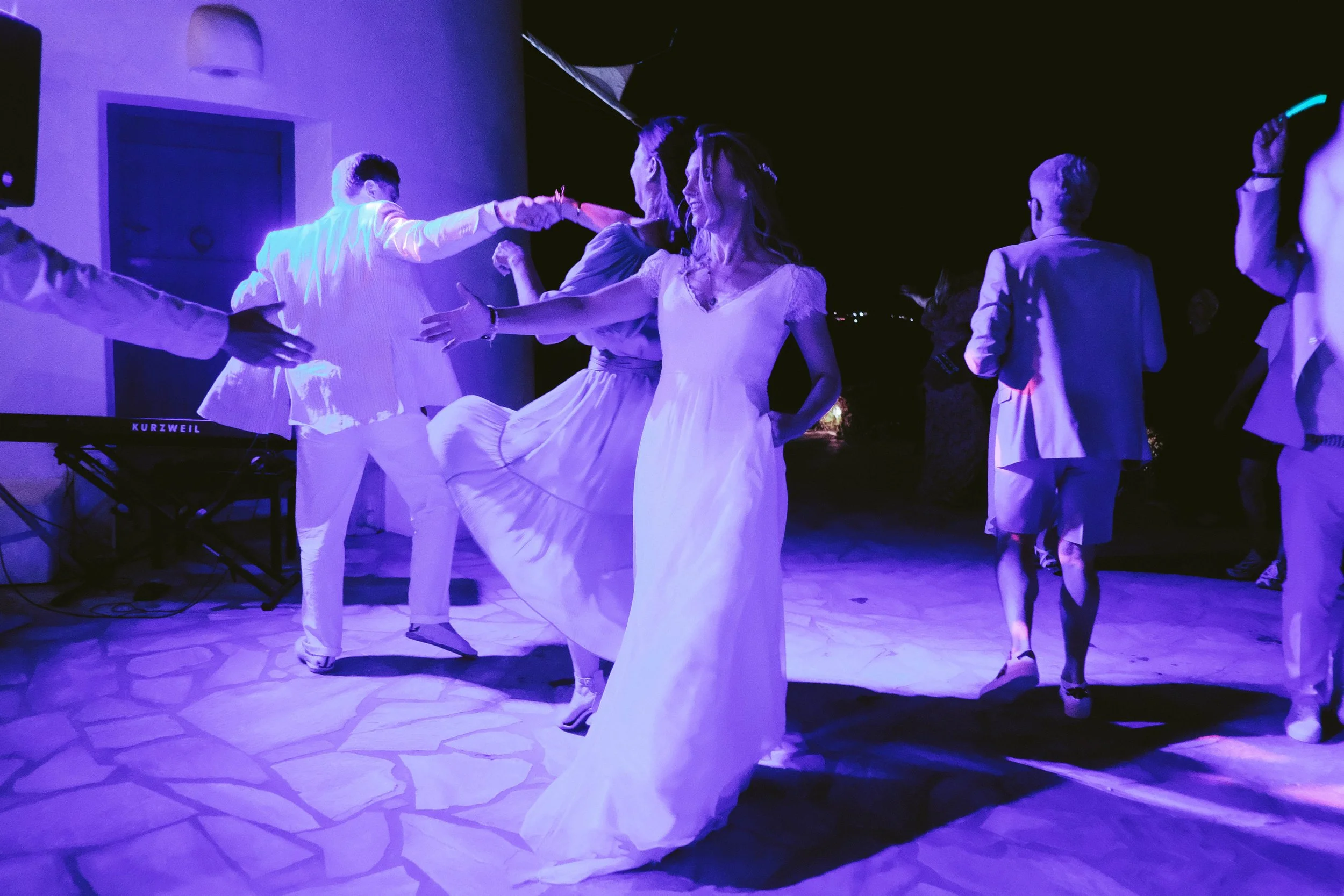 People dancing outdoor at night, illuminated by purple lights, with a woman in a white dress twirling.
