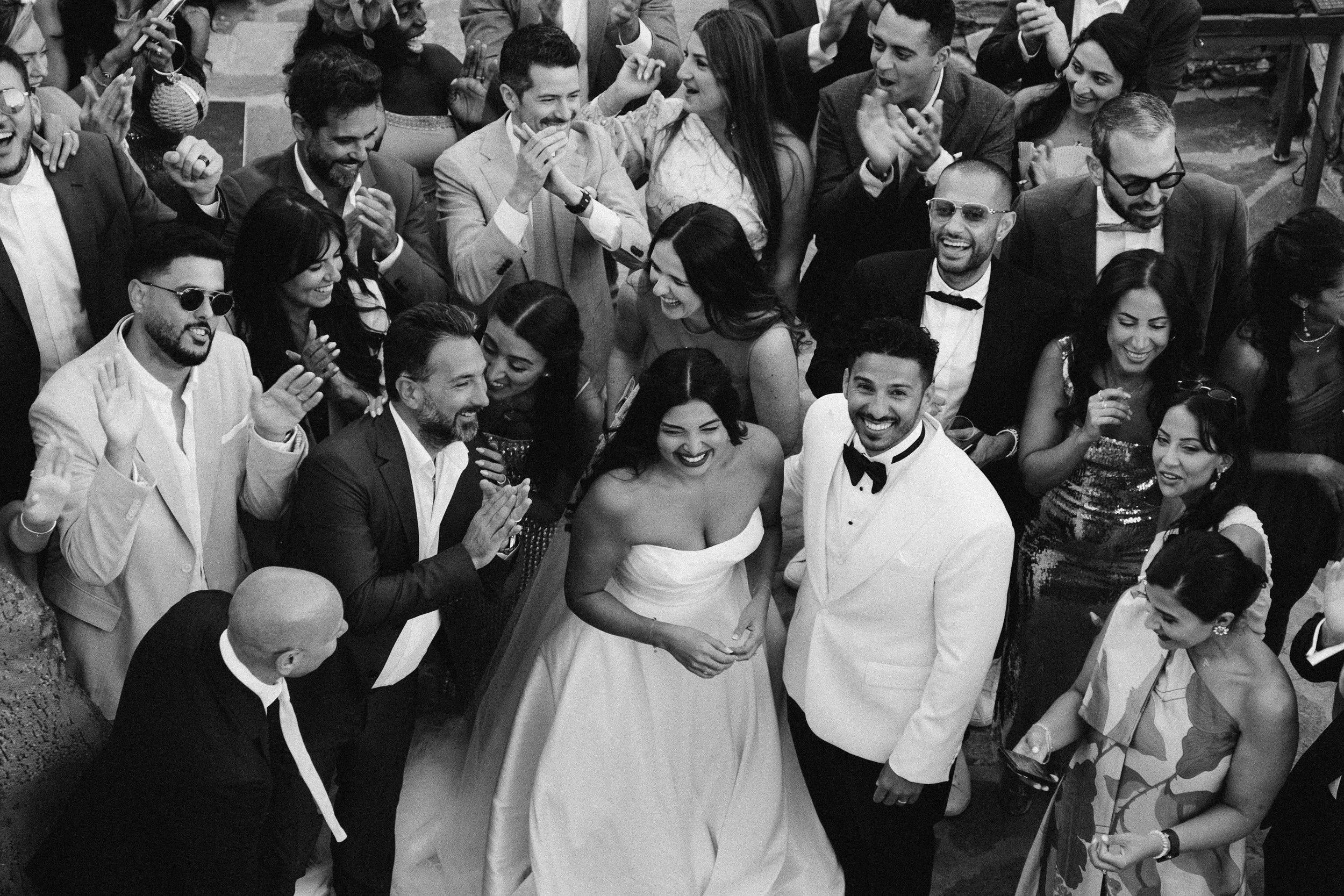 A black and white photo of a wedding celebration with a large group of people gathered around a smiling bride in a strapless wedding dress and a groom in a tuxedo. The guests are dressed in formal attire, laughing and clapping.