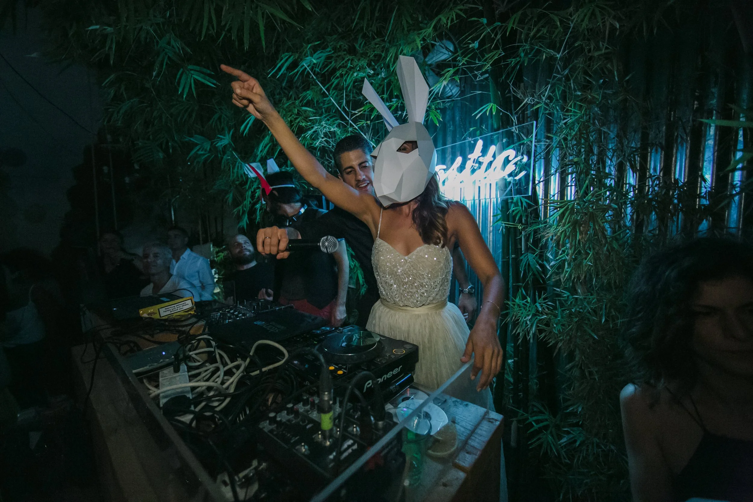 A woman wearing a white dress and a bunny mask holds a microphone and points upward while DJ equipment is in front of her at a party or nightclub, with a man standing behind her smiling and pointing.