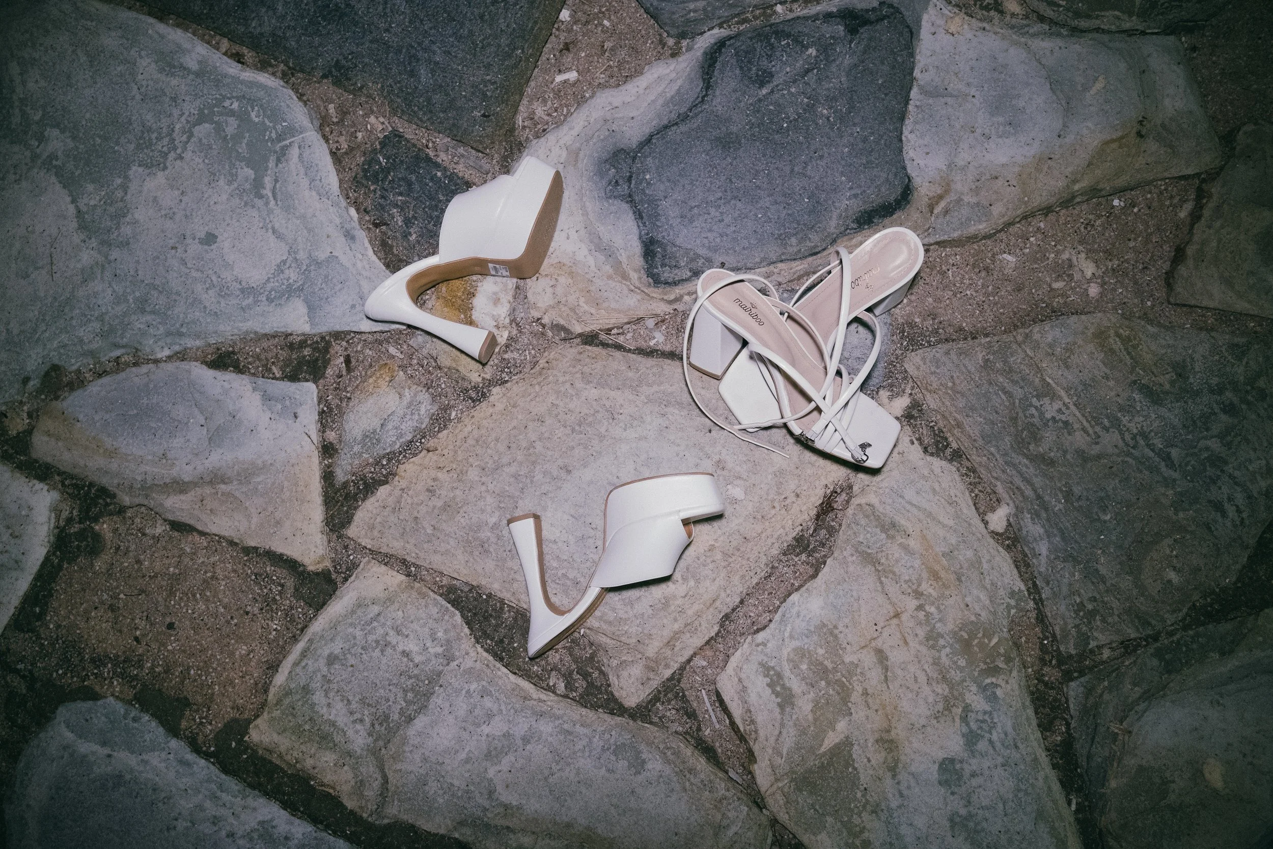 A pair of white high-heeled sandals on a stone pavement, with one shoe lying on its side and the other standing upright.