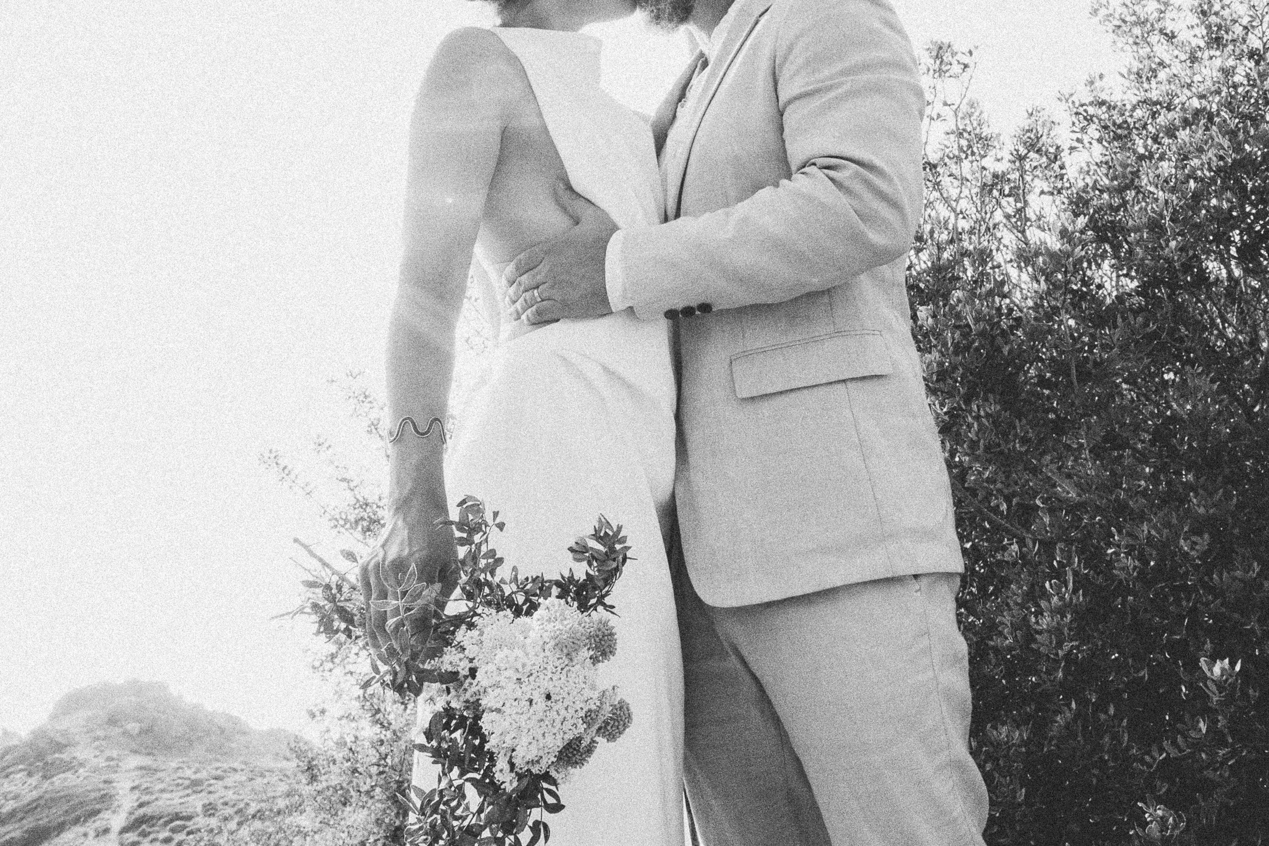 A black and white photo of a couple in wedding attire embracing outdoors, with the woman's hand holding a bouquet of flowers and the man's arm around her waist.