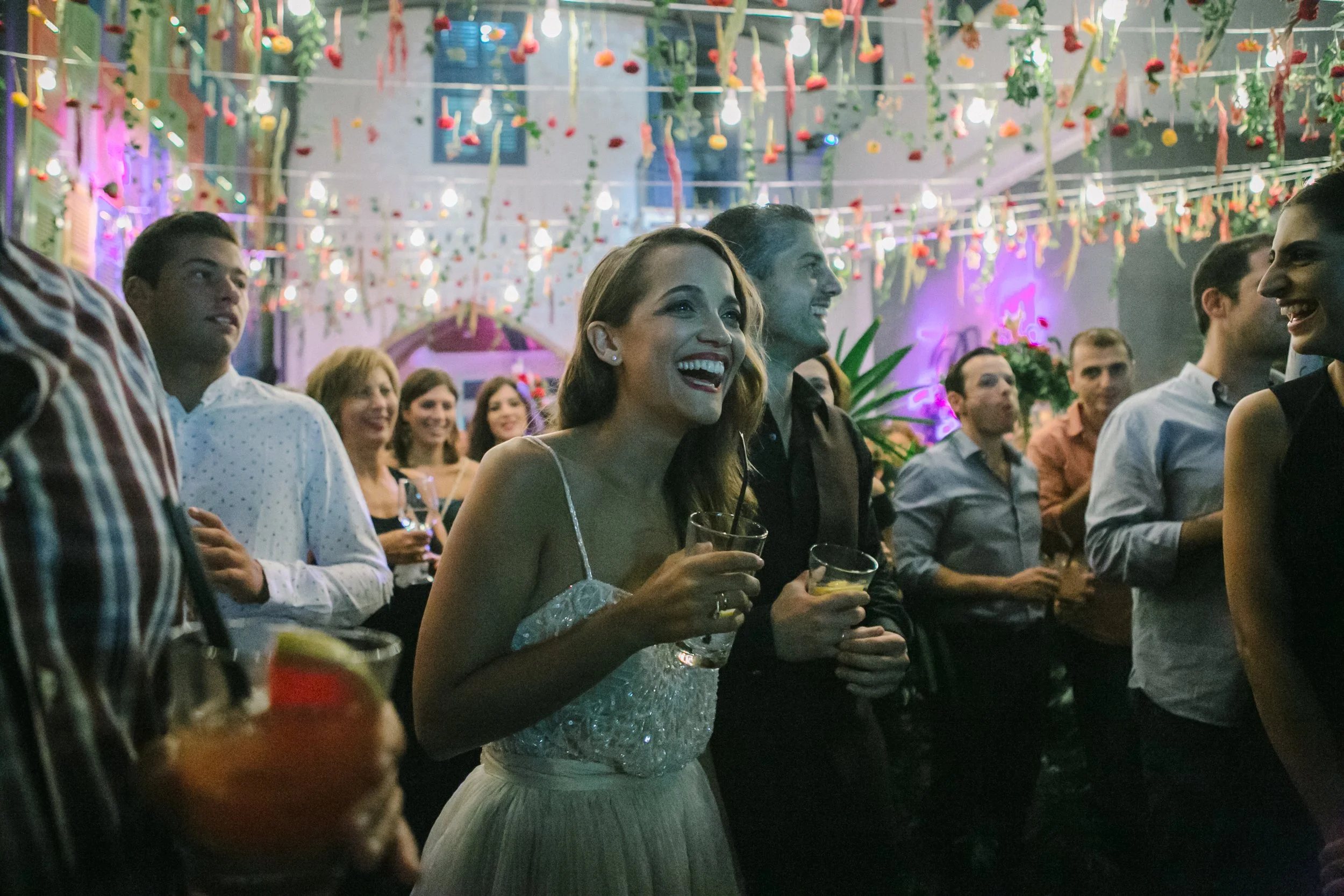 A group of people at a festive party or event, standing together, some holding drinks, and smiling. The setting is decorated with colorful streamers, hanging ornaments, and vibrant lighting.