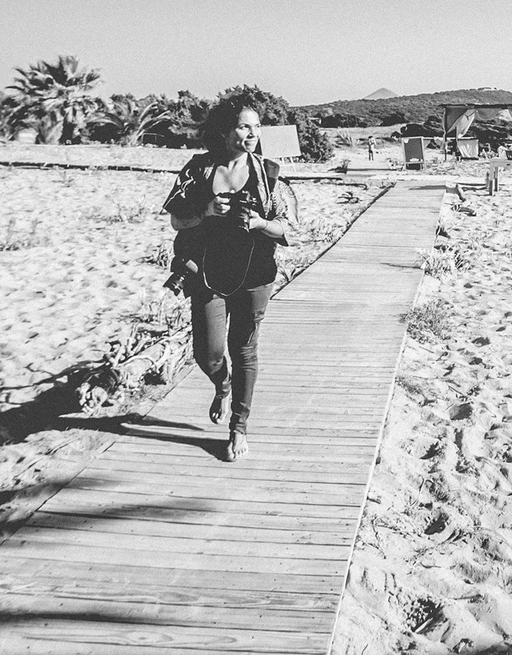 A woman with curly hair walking on a wooden walkway on a beach, holding a camera and smiling, with lounge chairs and trees in the background.