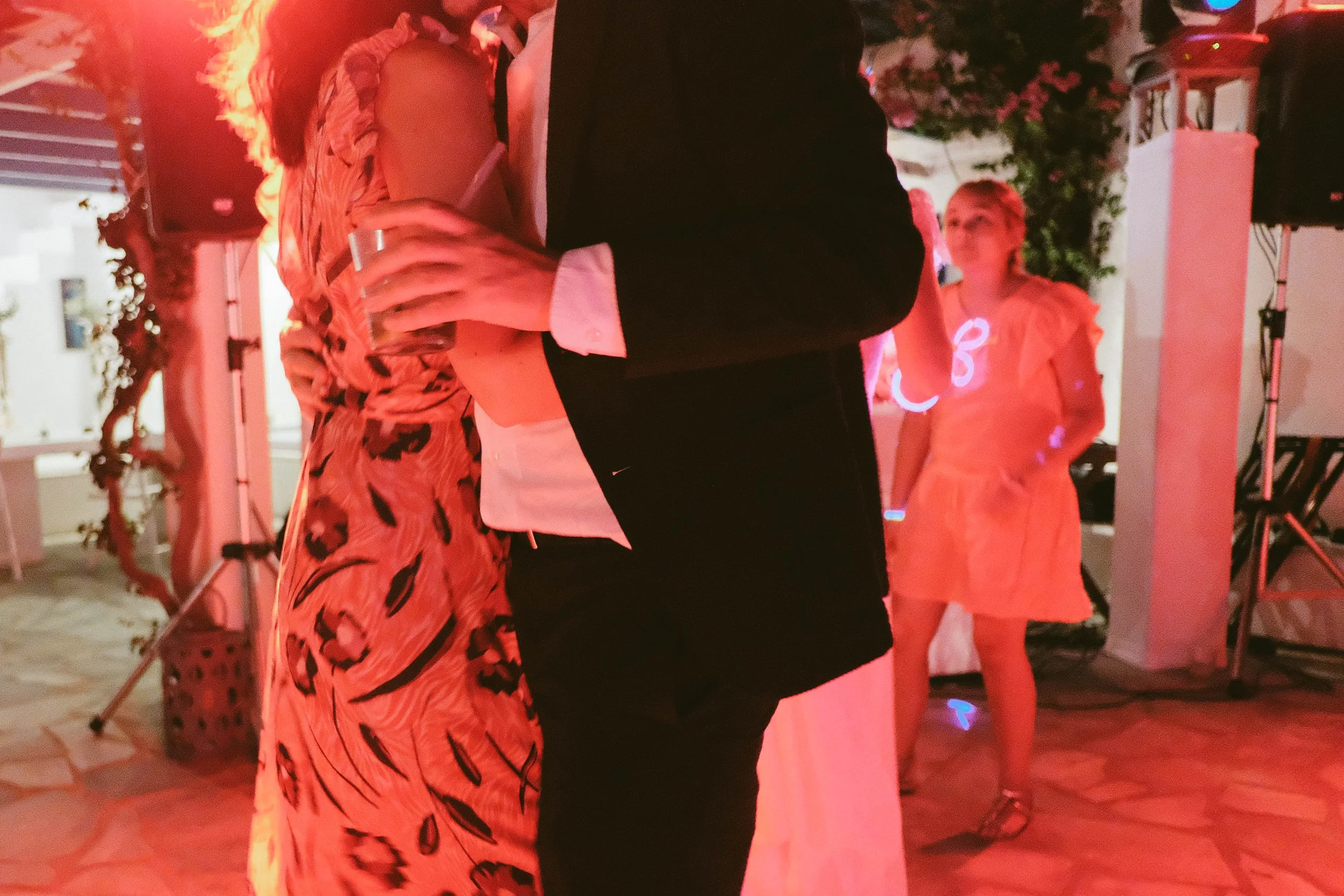 A couple dancing at a party, with the woman wearing a animal print dress and the man in a black jacket and white shirt, and a woman in the background wearing a pink dress.