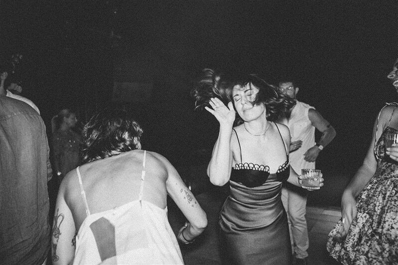 People dancing at a party, woman in a satin dress with her hair swinging, others holding drinks in a dark, lively setting.