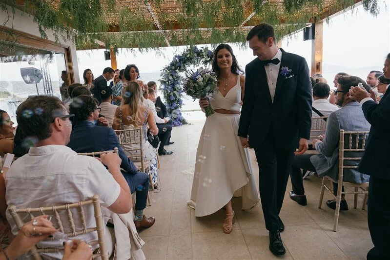 A bride and groom walking down the aisle at a wedding ceremony with seated guests on both sides and a floral arch in the background.