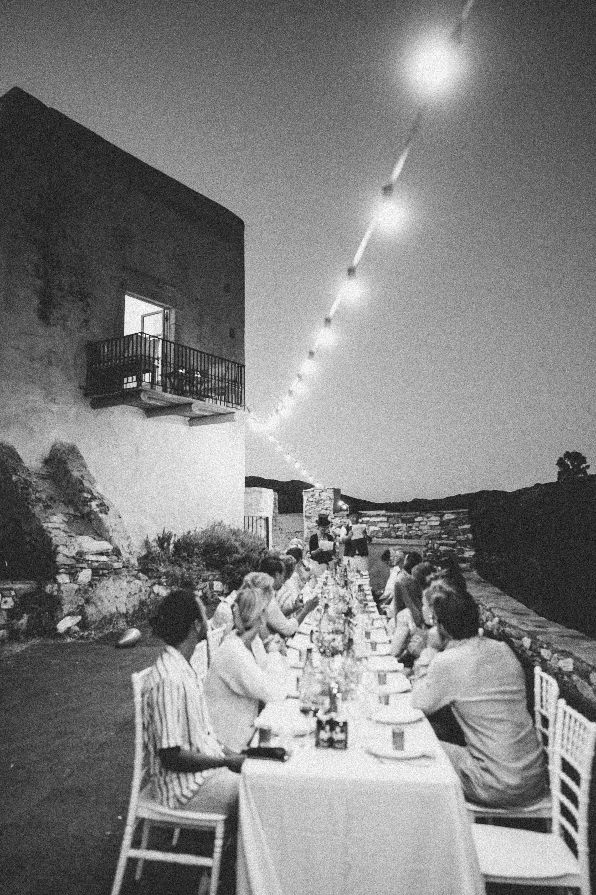 People enjoying dinner at an outdoor long table during evening, with string lights overhead, outdoor setting with building and landscape in background.