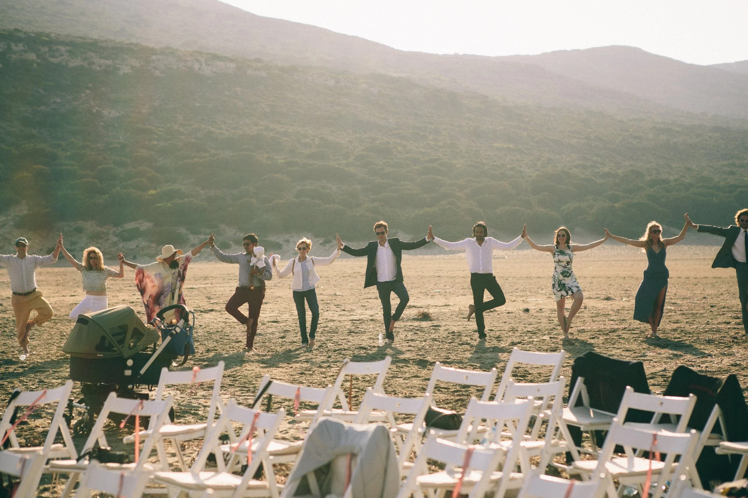 A group of people dancing on a beach, holding hands, with chairs and a stroller in the foreground and mountains in the background.