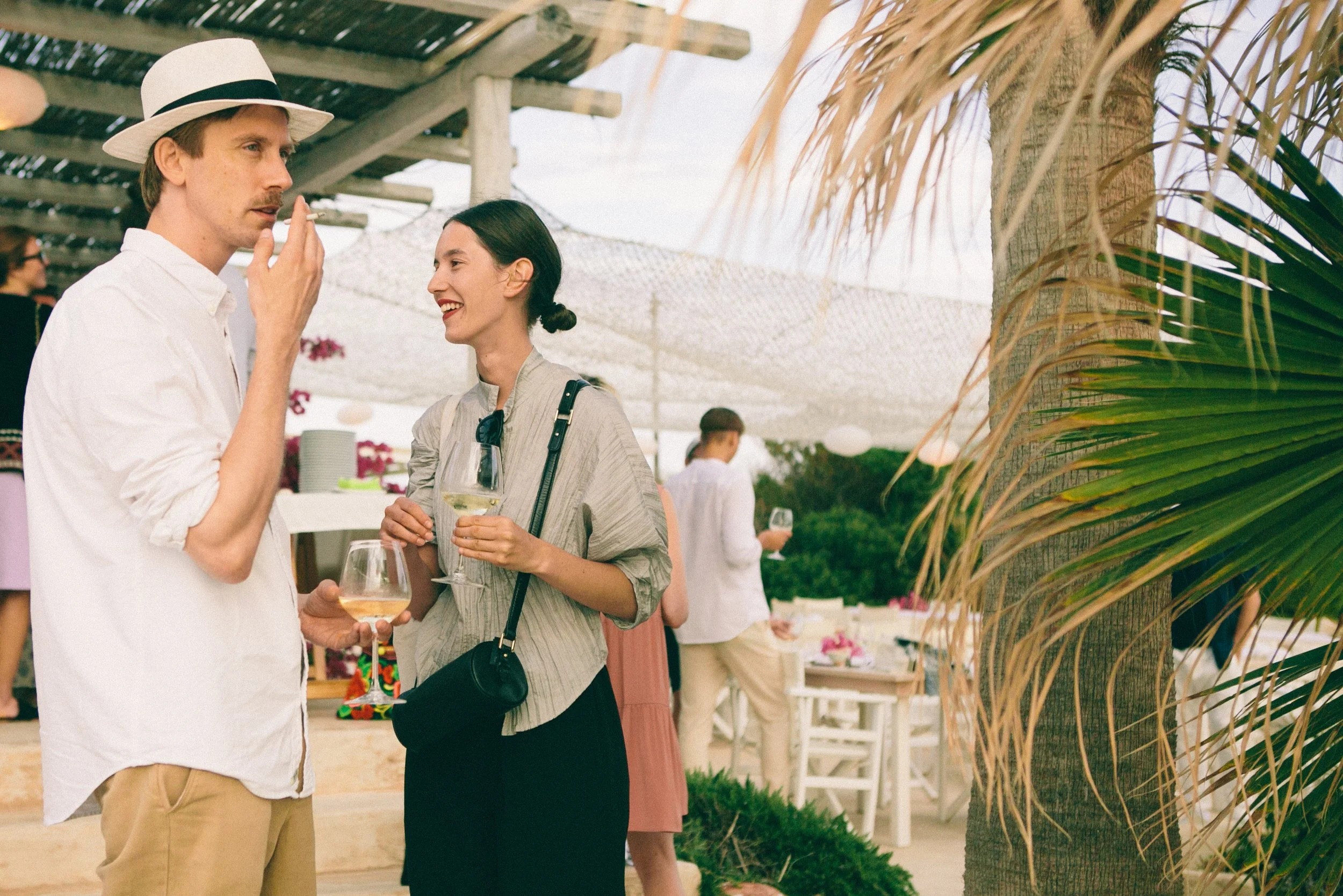 Two people, a man and a woman, are attending an outdoor event on a beach or patio, holding wine glasses. The man is wearing a white shirt and a straw hat, and is holding a cigarette, while the woman is wearing a gray striped shirt and has sunglasses 