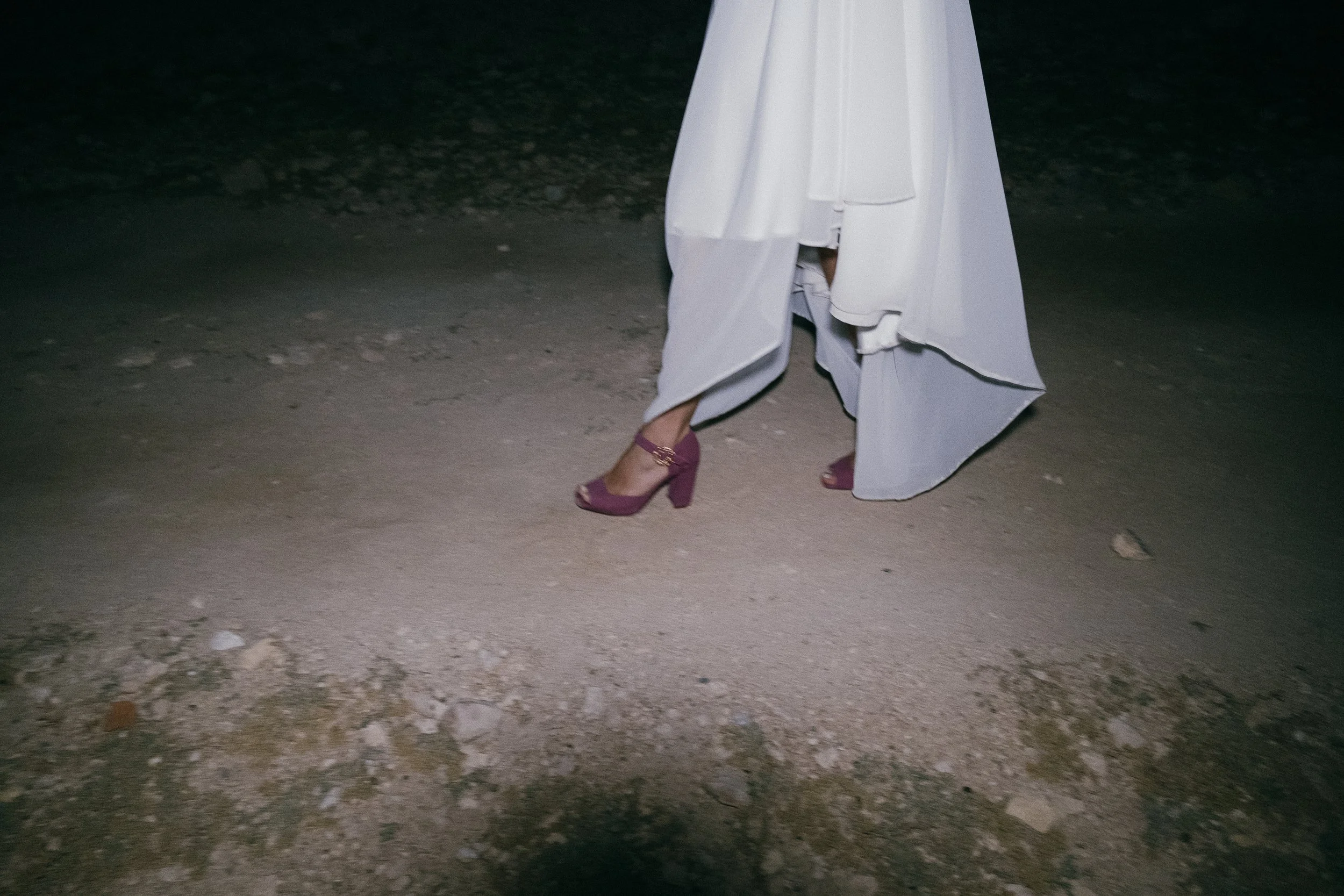 Person wearing high-heeled shoes and a white dress walking on a dirt road at night.