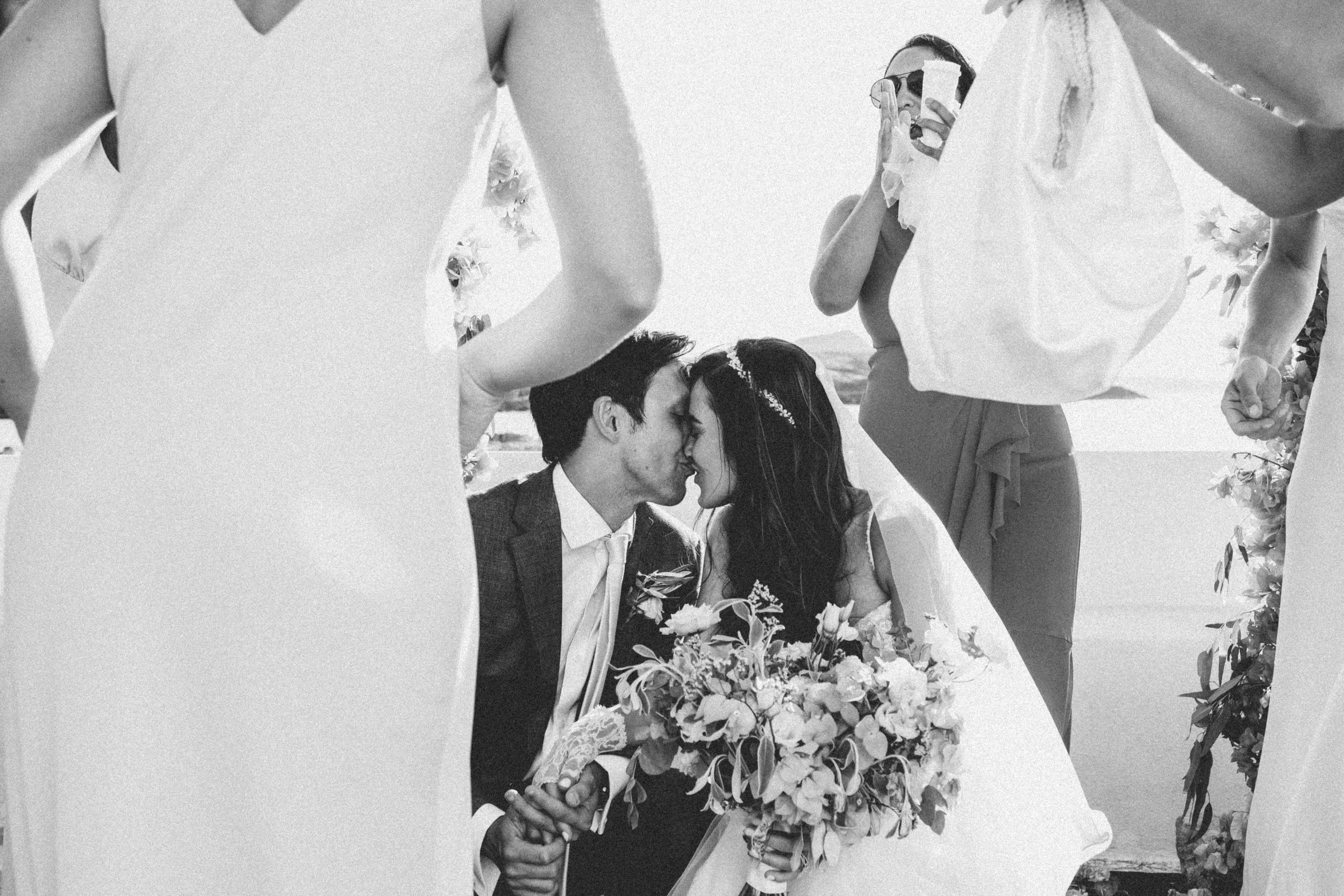 A black and white photograph of a wedding ceremony with a couple kissing, surrounded by their wedding party. The couple is seated, holding hands, with the bride holding a bouquet of flowers. The bride and groom are facing each other with their forehe