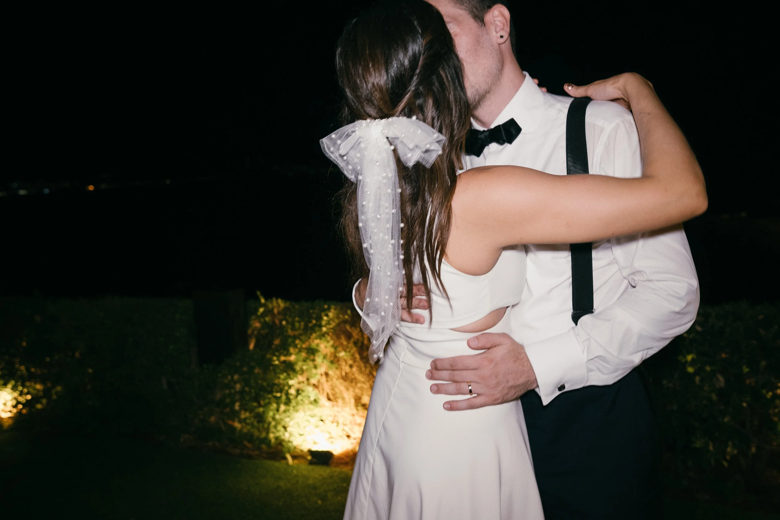 Couple kissing at night, dressed in wedding attire, outdoors with illuminated bushes in background.