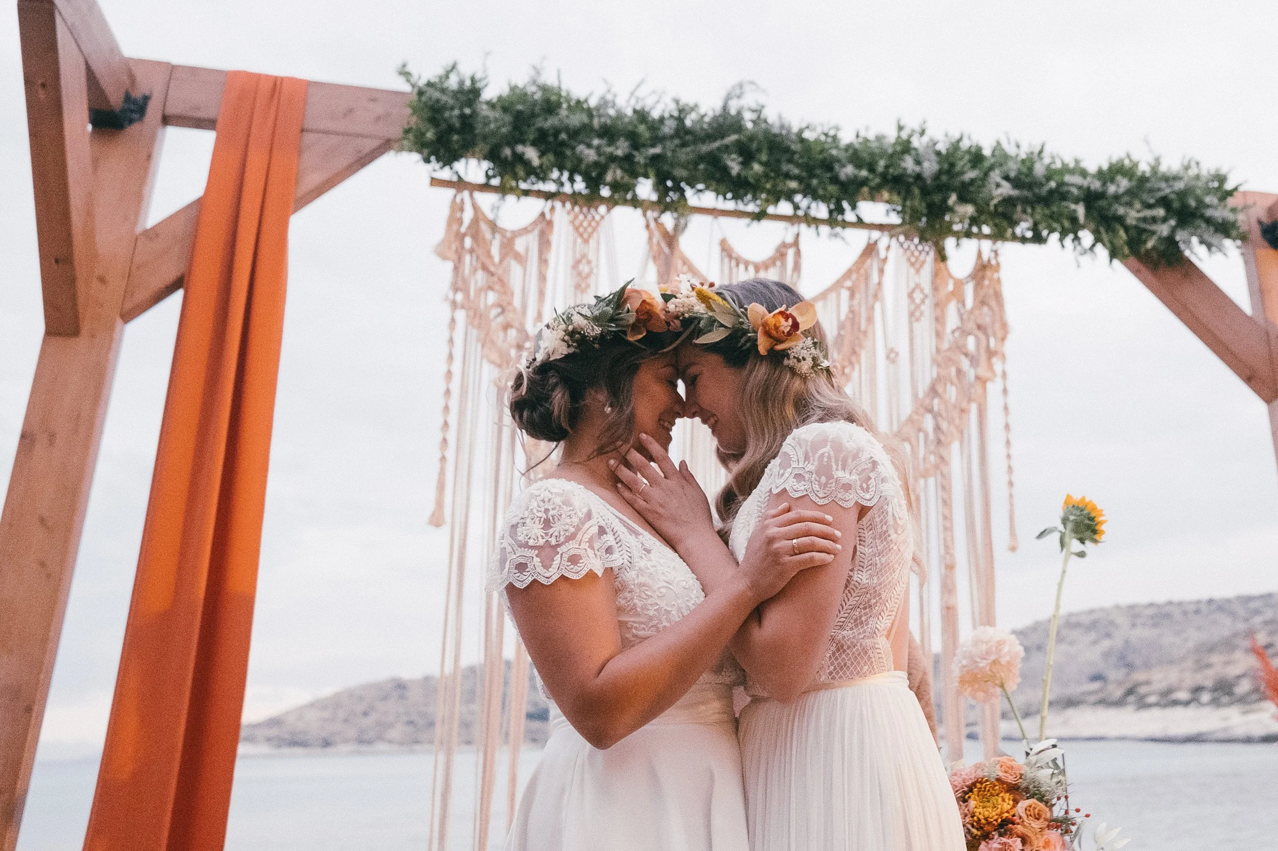 Two women in wedding dresses sharing a tender moment under a decorated arch on a beach.