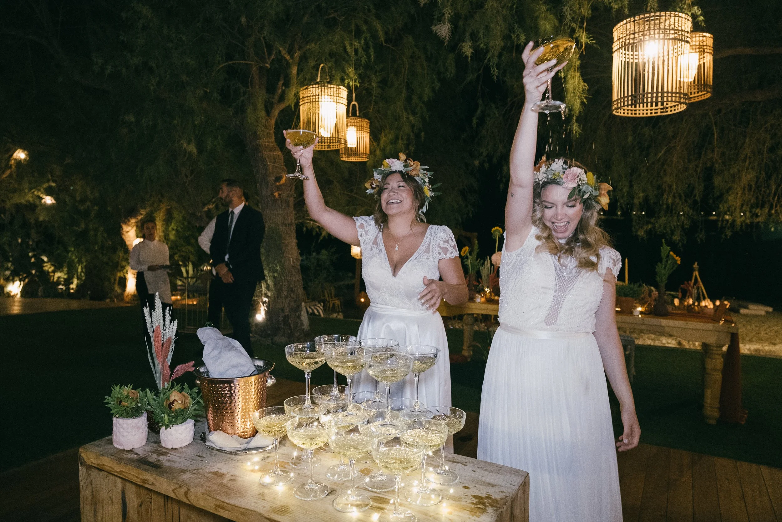 Two women in white dresses with floral crowns toasting with champagne glasses at a celebration outdoors at night, with a table of champagne glasses, flowers, and decorative lights.