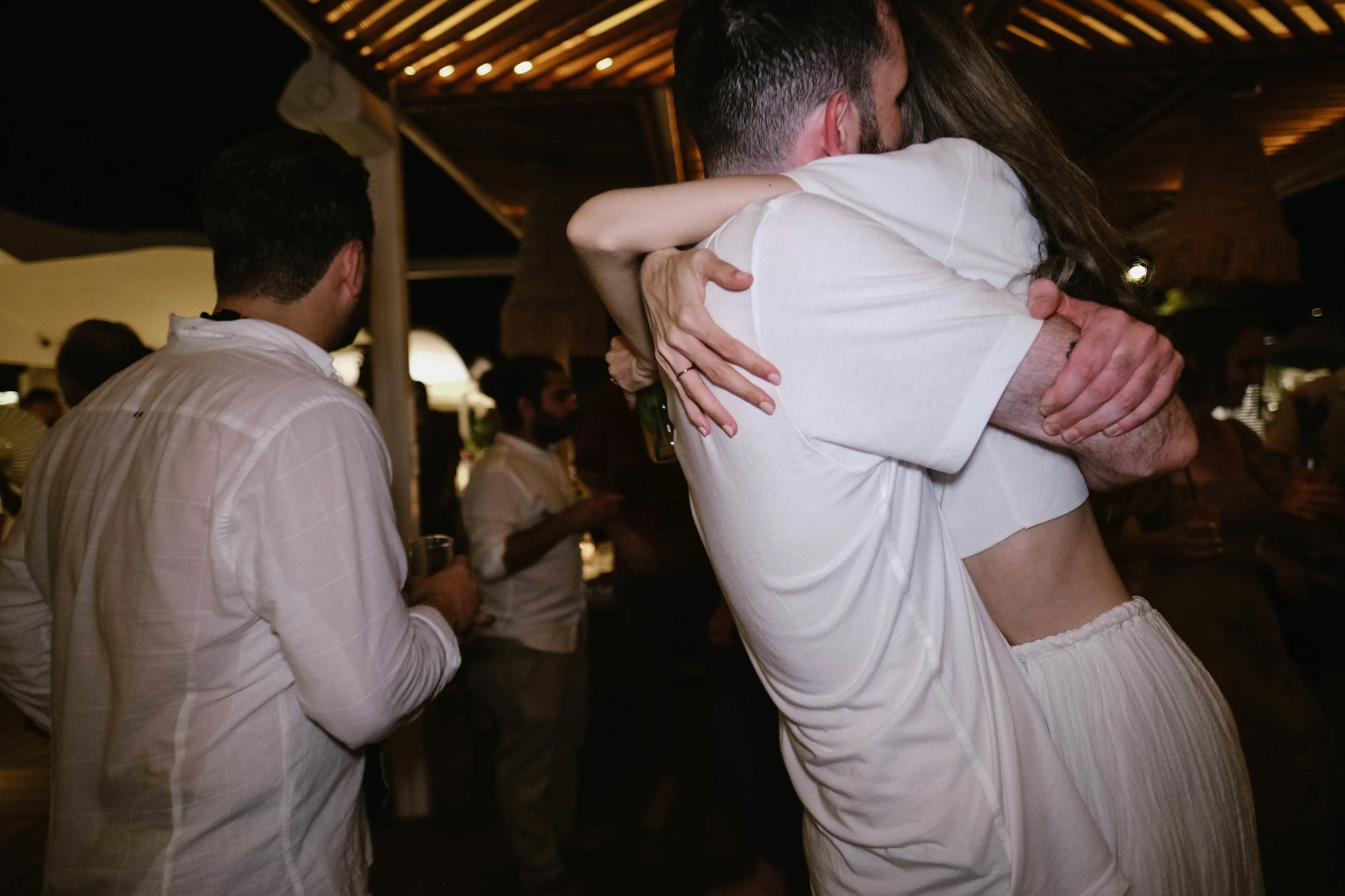 People hugging at a social event, dressed in light-colored clothing, with a man in the foreground holding a drink, engaged in conversations in a dimly lit outdoor setting.