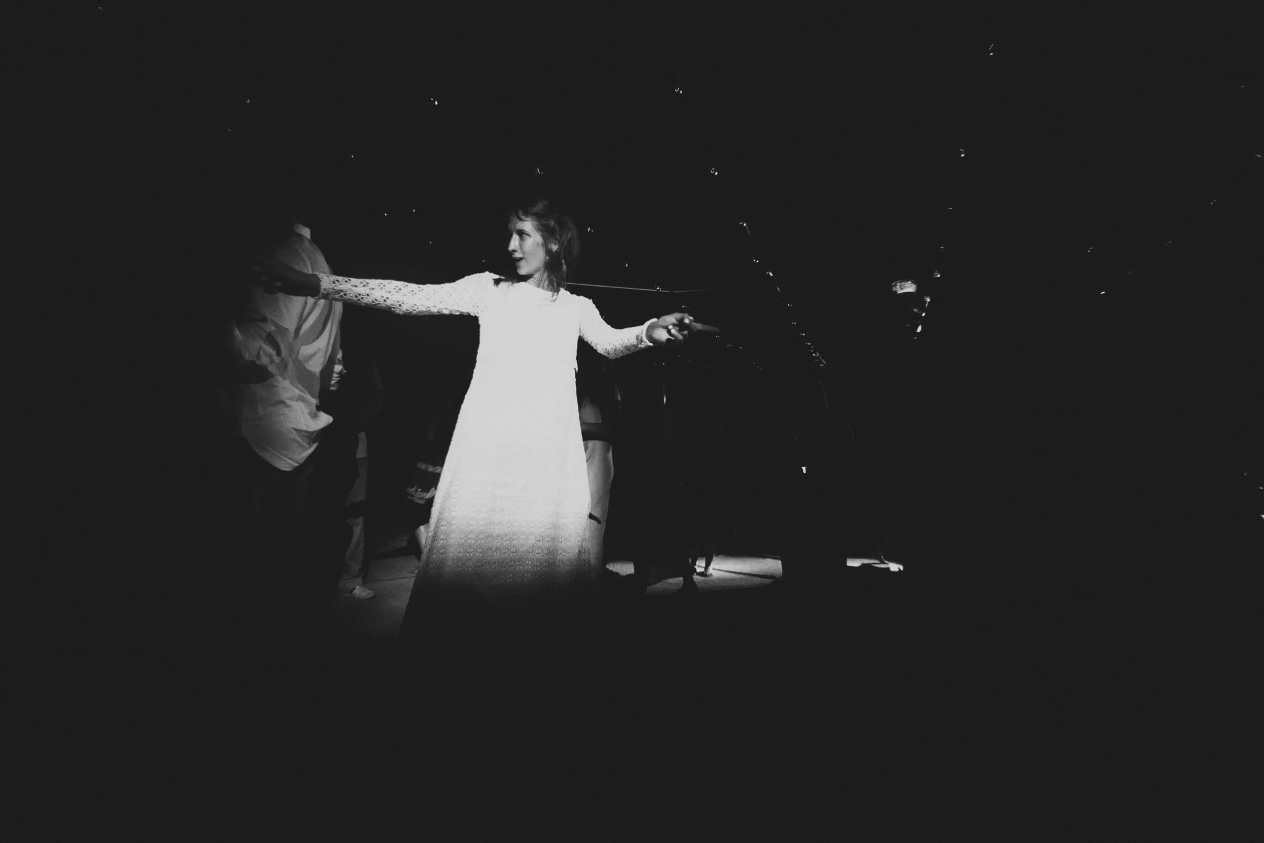 A woman in a long white dress reaching out with both arms, standing in the dark with some people faintly visible in the background.
