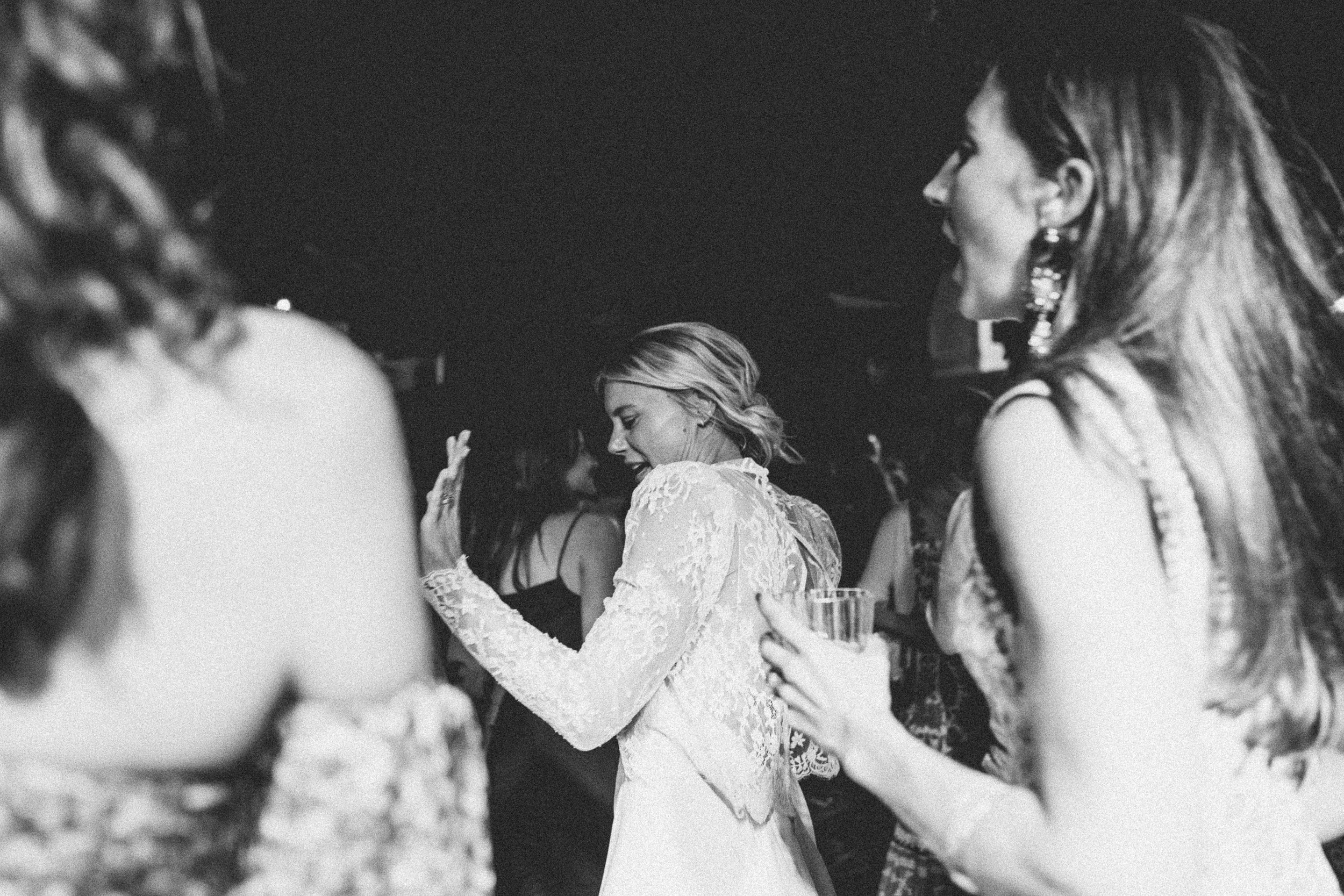 Black and white photo of women dancing at a party or wedding, with a woman in a lace dress smiling and raising her hand.