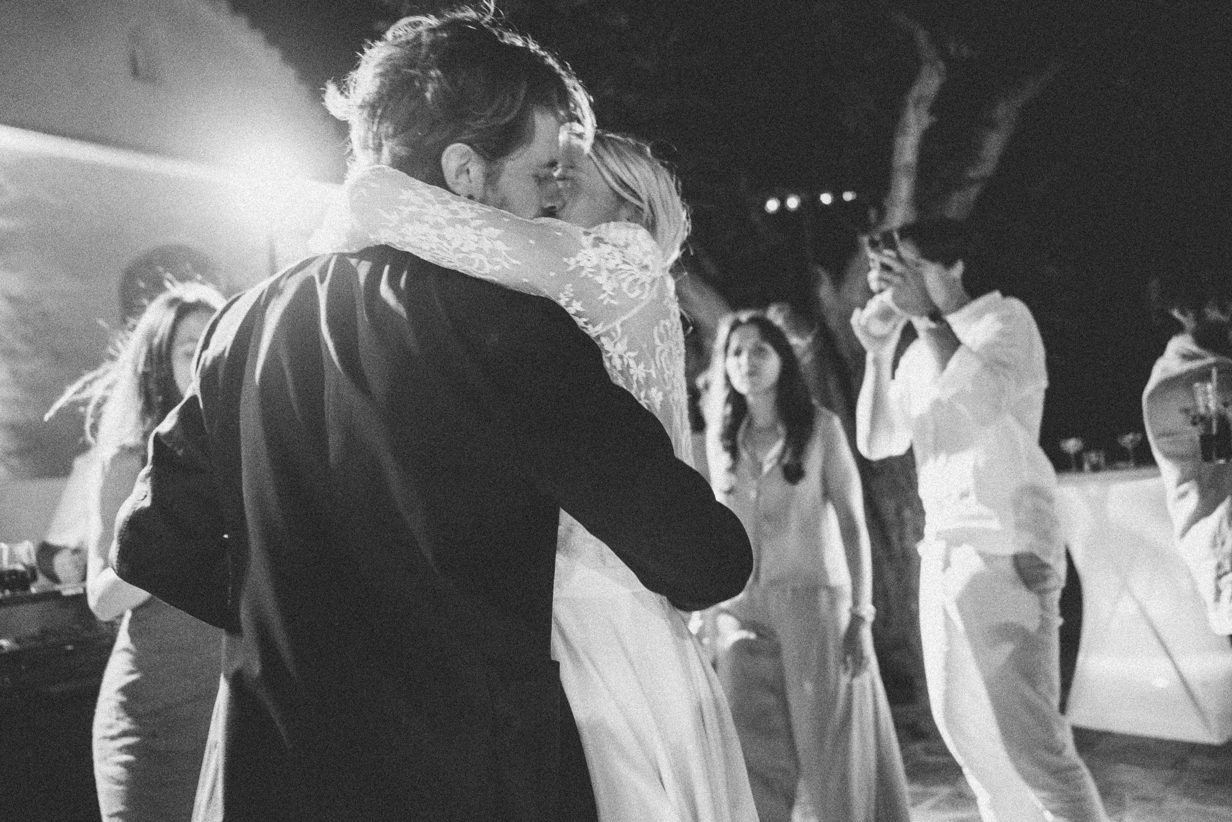 A couple dancing closely at an outdoor event at night, surrounded by guests, with a tree and string lights in the background