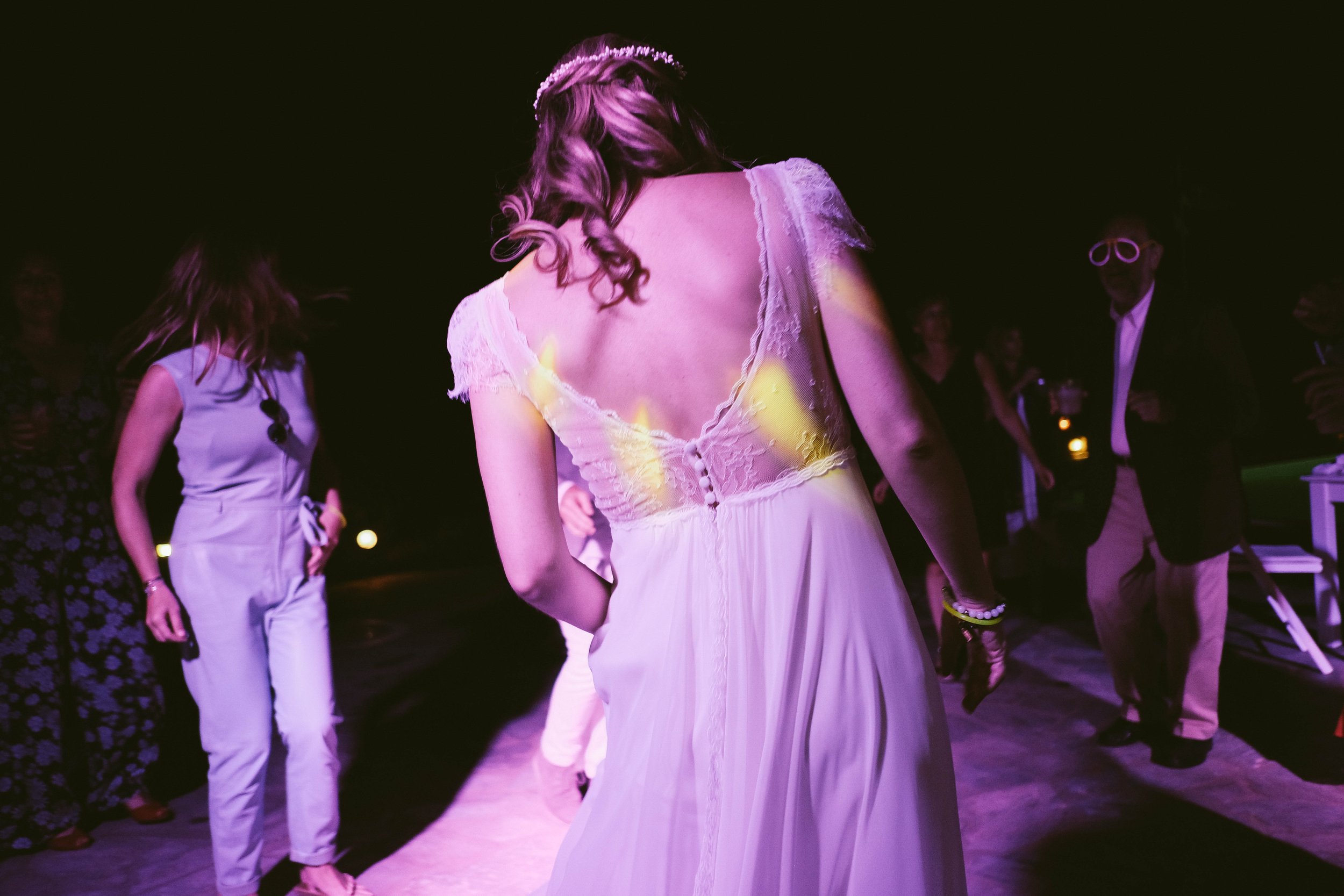 Woman dancing at a party or wedding reception wearing a white dress with lace back, others dancing in the background at night