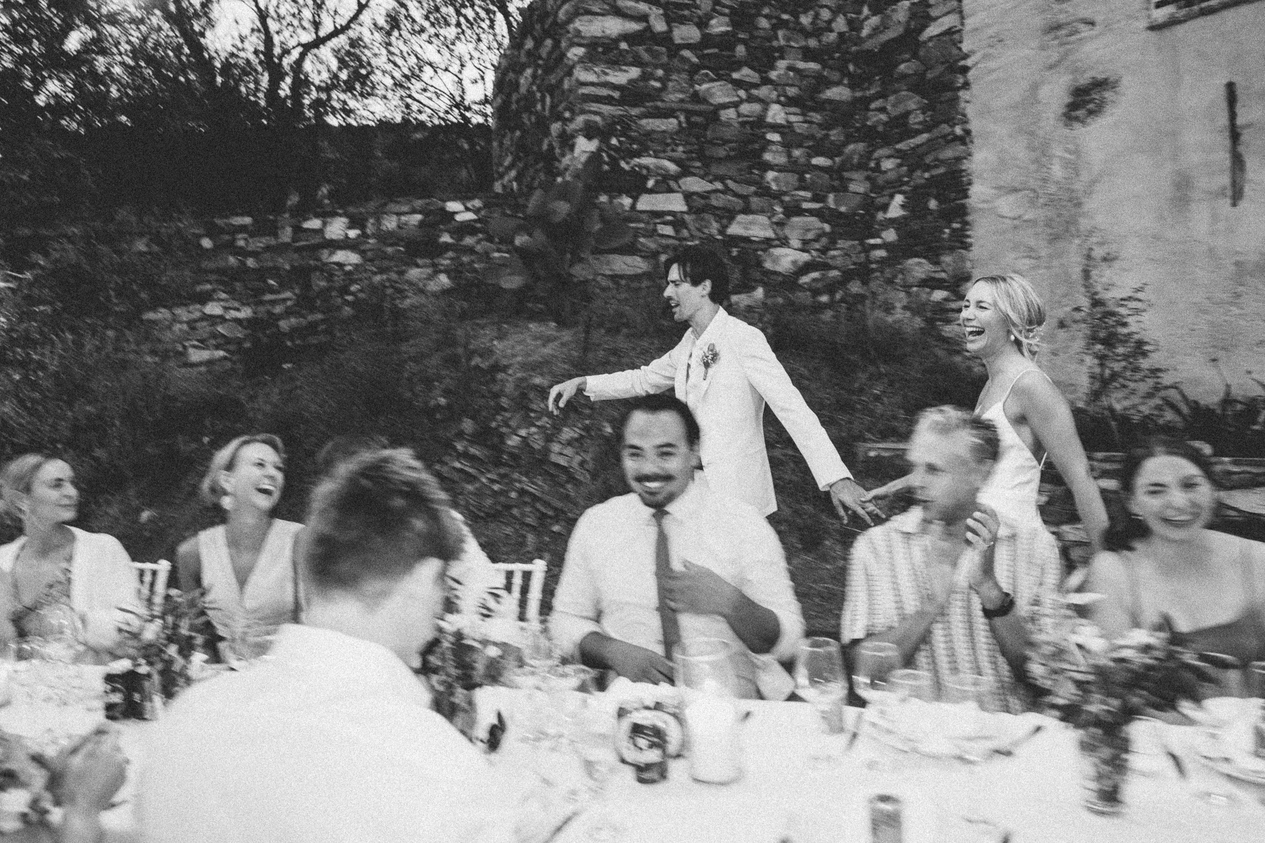 A black and white photo of a joyful outdoor gathering with seven people sitting at a table, and two people standing and holding hands behind the table. The people are smiling and laughing, with a stone wall and trees in the background.