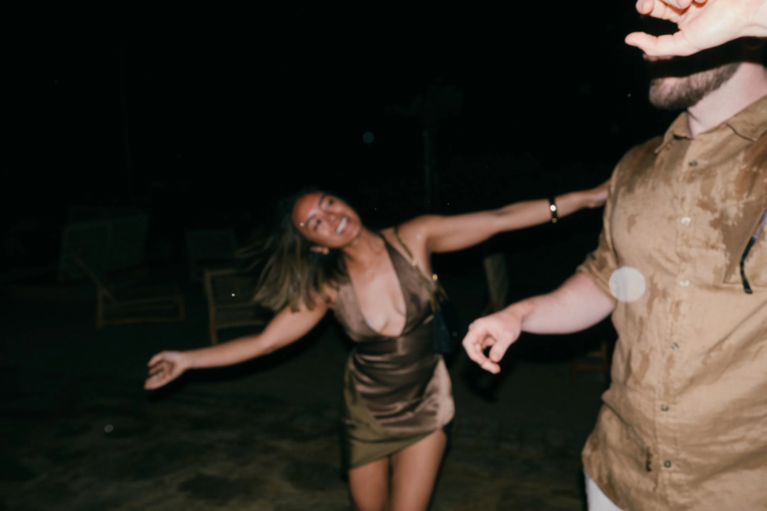 A woman dancing happily at night with outstretched arms, next to a man who is partially visible in the foreground.