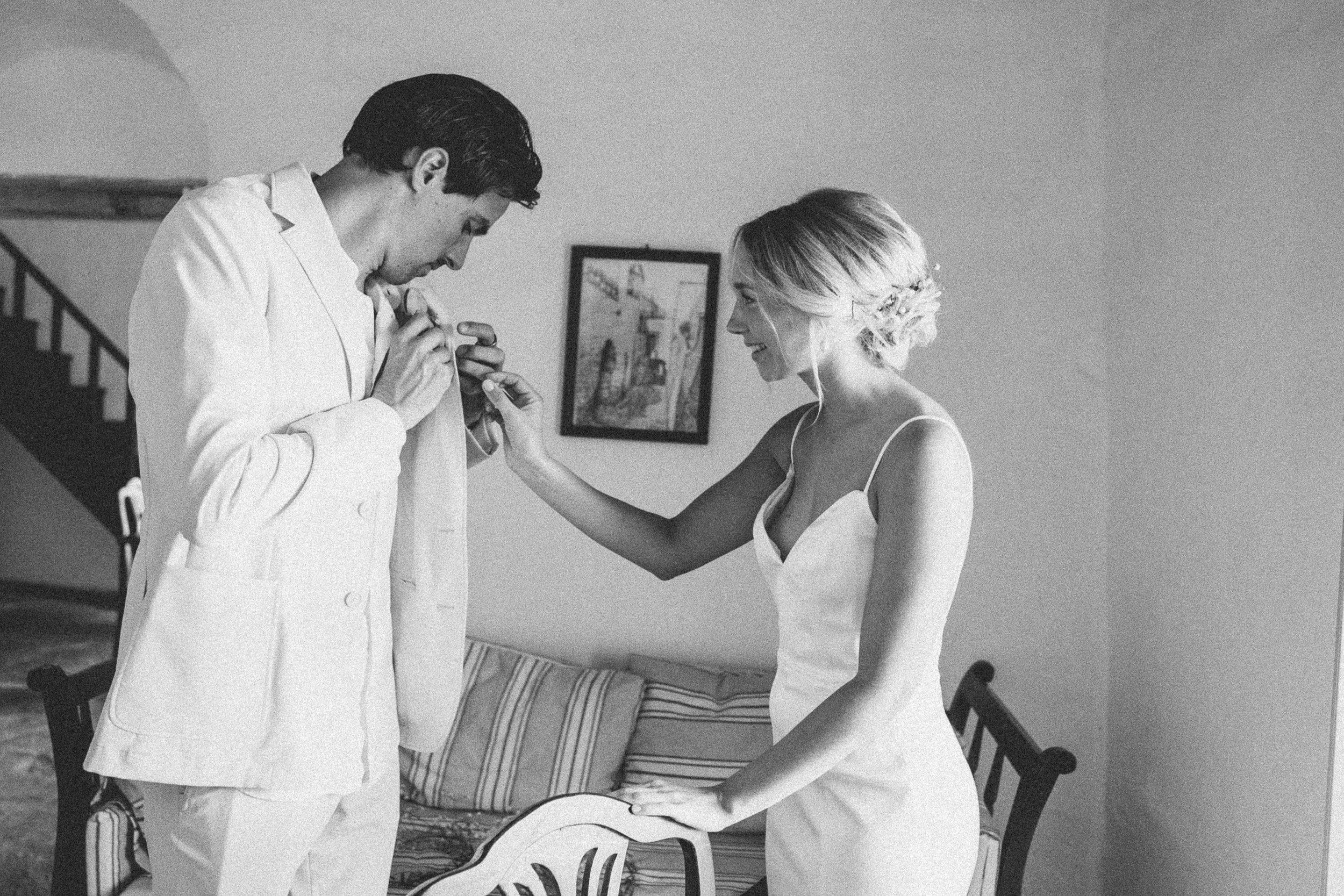 A bride with a floral hair accessory and a groom in a light-colored suit, smiling indoors, with a couch, a chair, and framed artwork on the wall in the background.