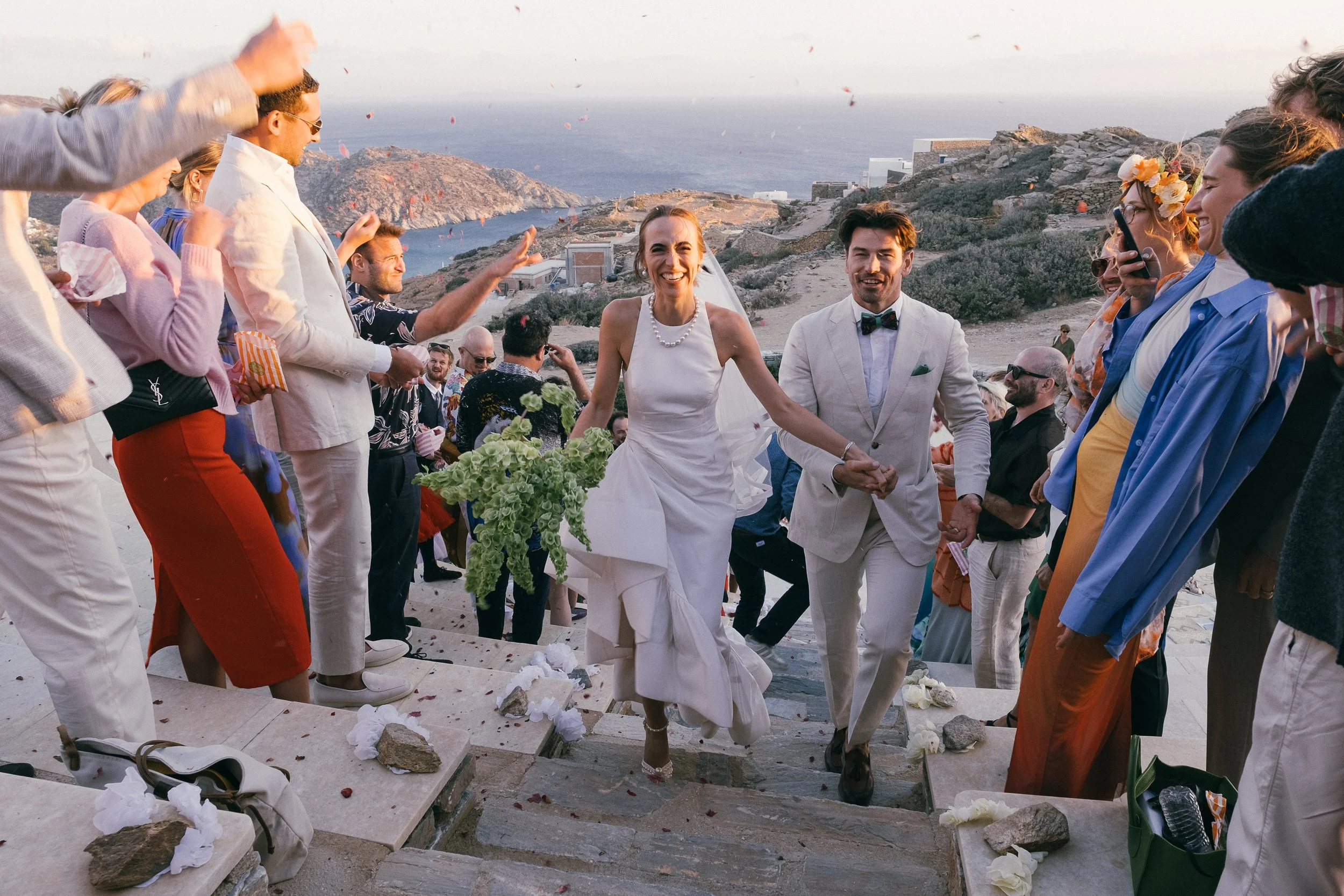Bride and groom walking down the steps of a wedding venue with a scenic ocean view, surrounded by guests celebrating.