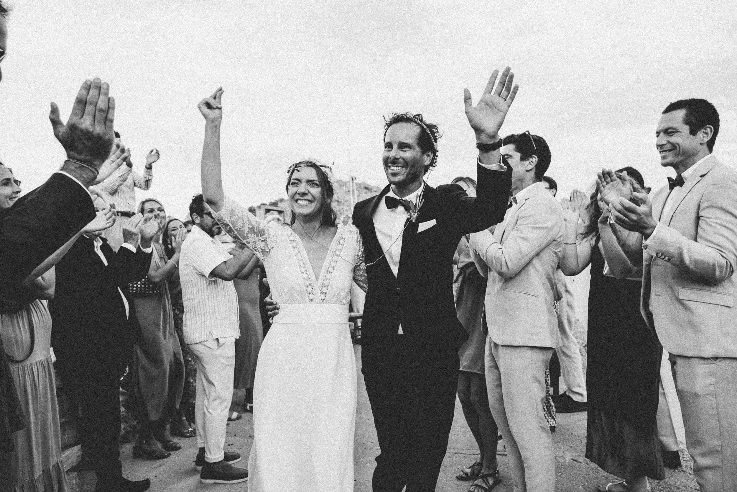 A wedding celebration with the bride and groom smiling and waving, surrounded by guests clapping and cheering outdoors.