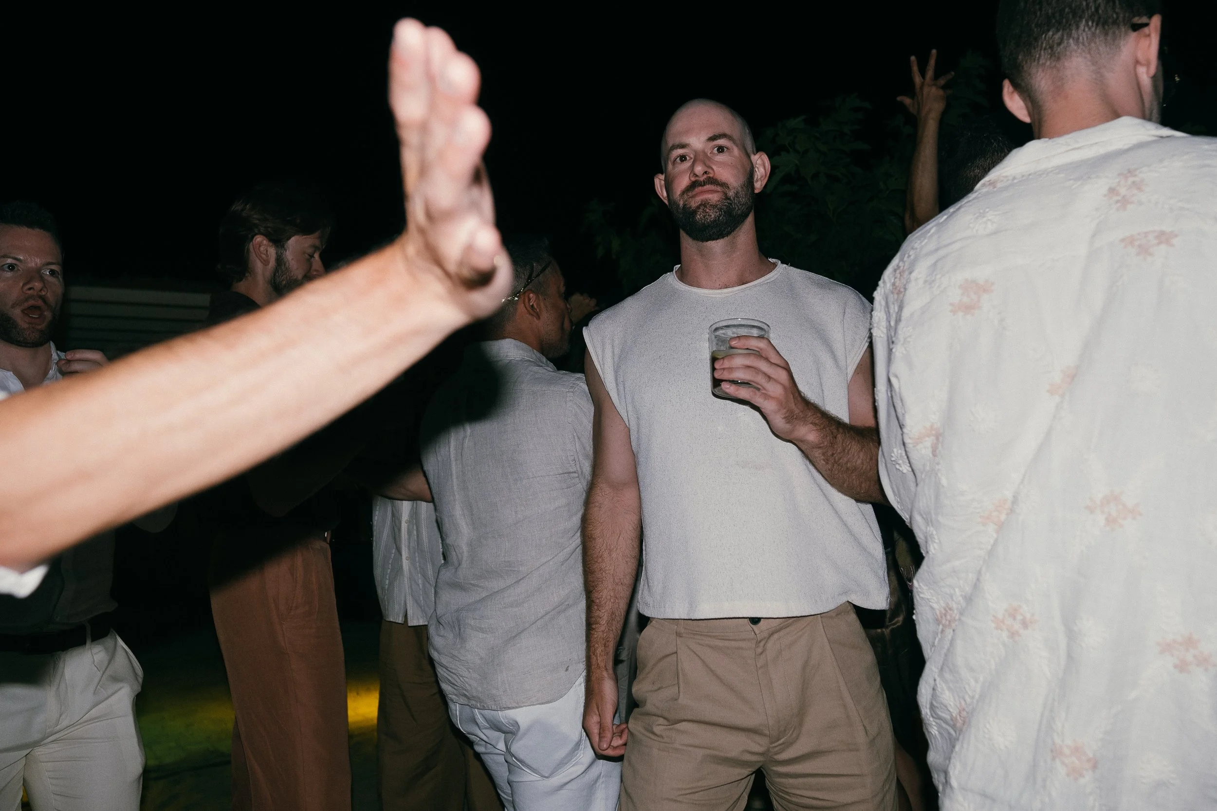 Group of men socializing outdoors at night, one man in the center holds a drink and looks at the camera, others engage in conversation.